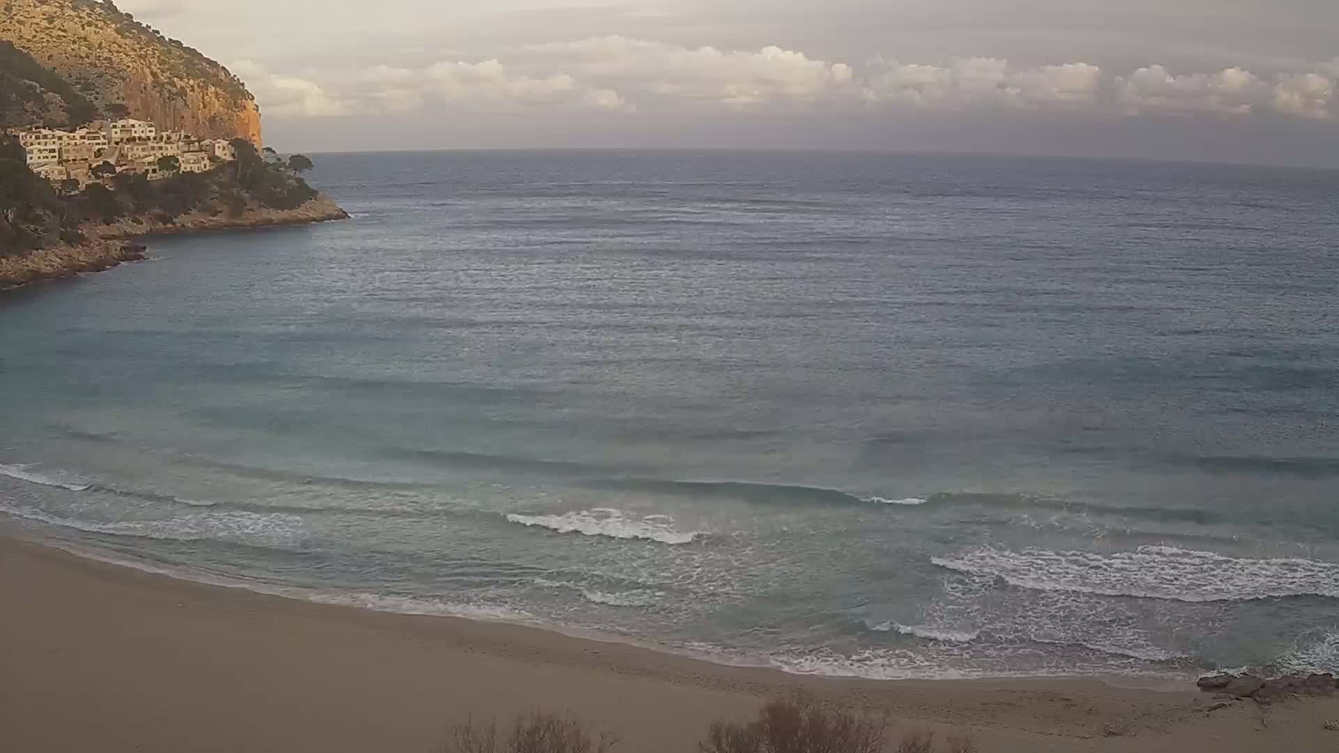 A sandy beach with small waves laps the shore under a cloudy sky,  with buildings nestled on a rocky cliff overlooking the ocean.