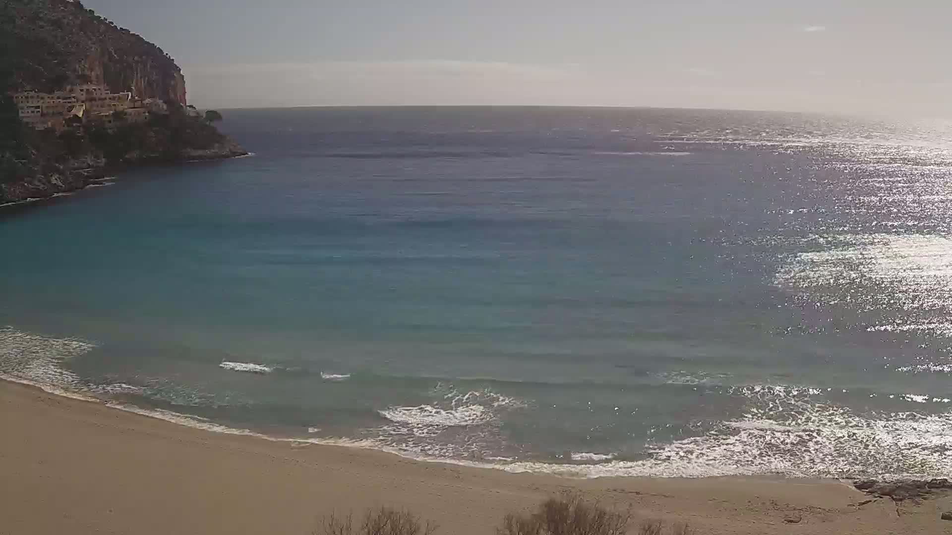 A sandy beach curves into a calm, teal ocean under a bright, sunny sky, with a cliffside and buildings visible in the distance.