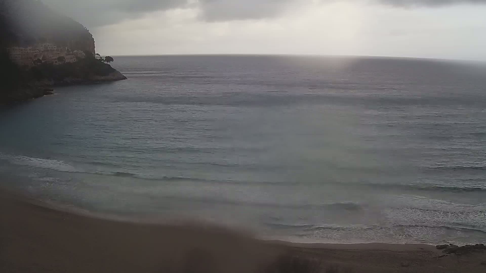 A sandy beach meets a calm, gray sea under an overcast sky, with a rocky cliff and buildings visible in the background.