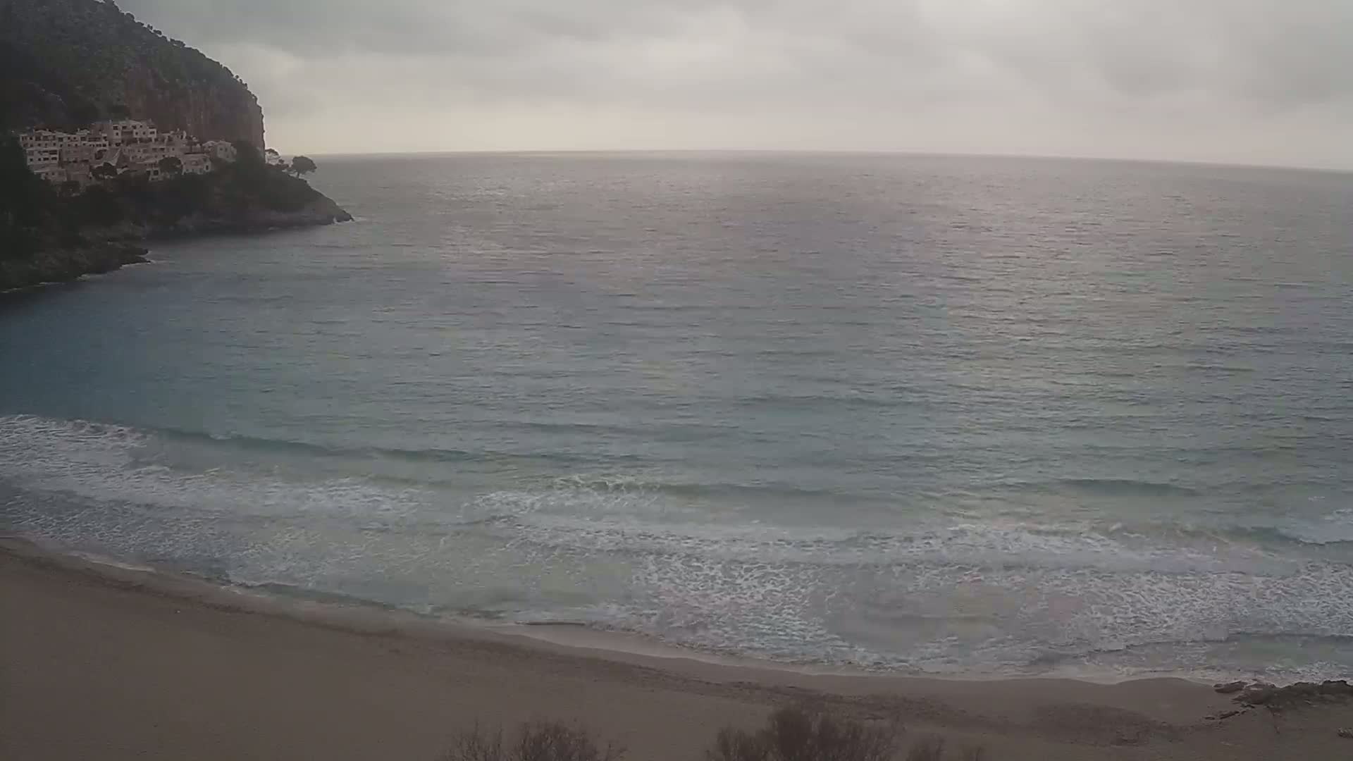 A sandy beach with gentle waves rolls onto the shore under an overcast sky, next to a rocky cliff with buildings on top.