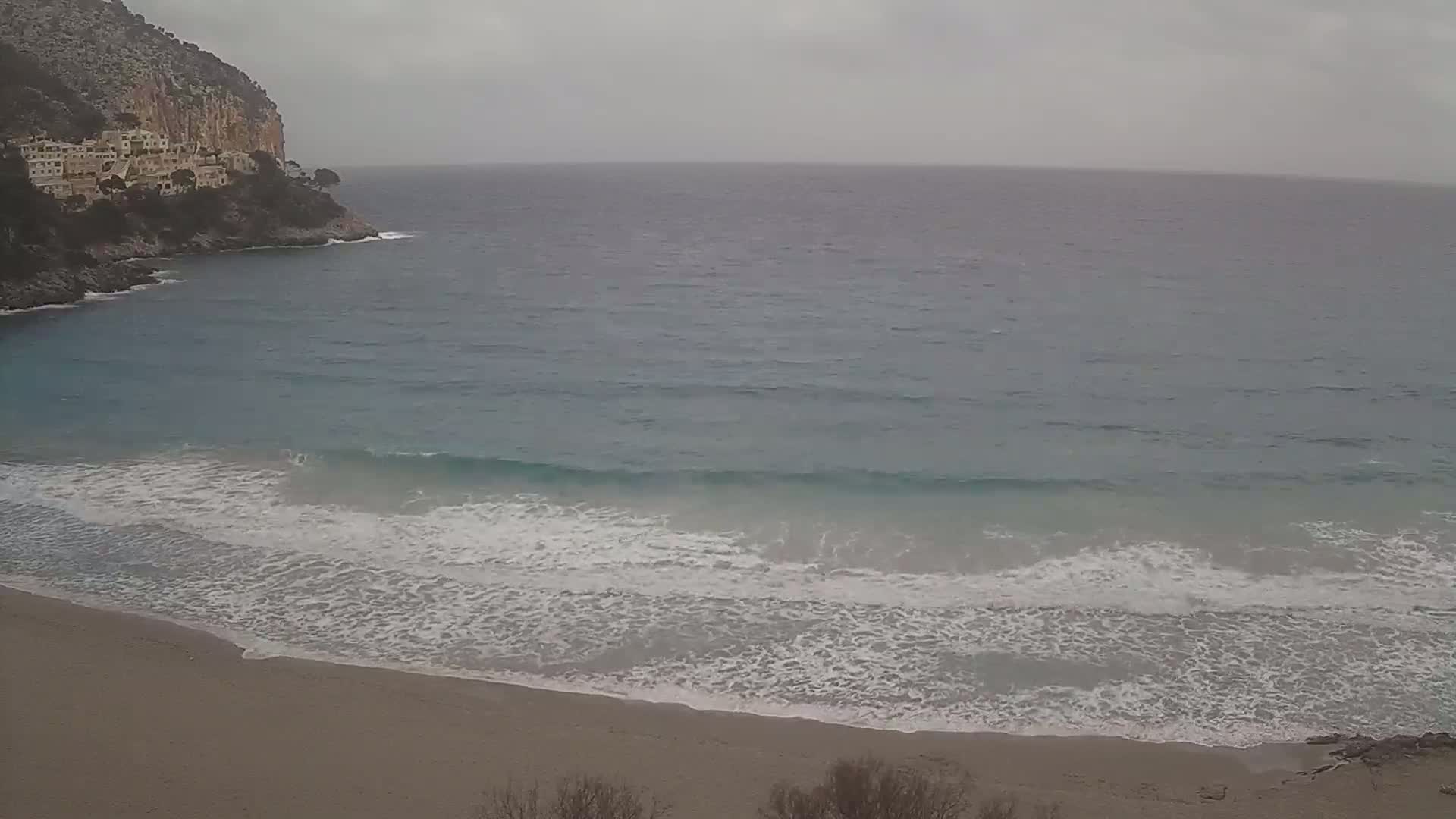 A sandy beach with small waves rolls onto the shore under an overcast sky, with buildings nestled on a cliff overlooking the ocean.