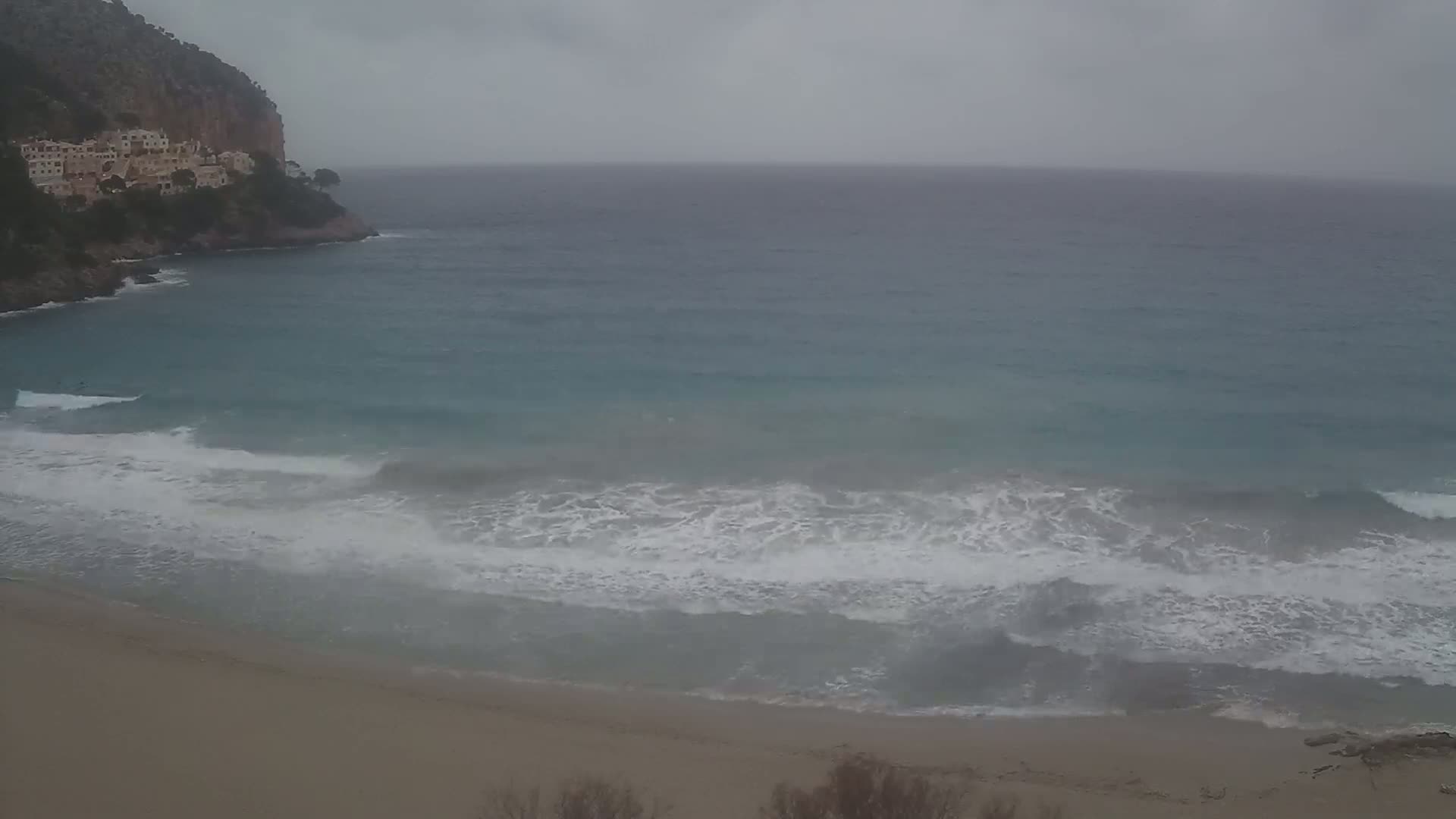 A sandy beach with moderate waves rolls onto the shore beneath a cloudy sky near a cliffside village.