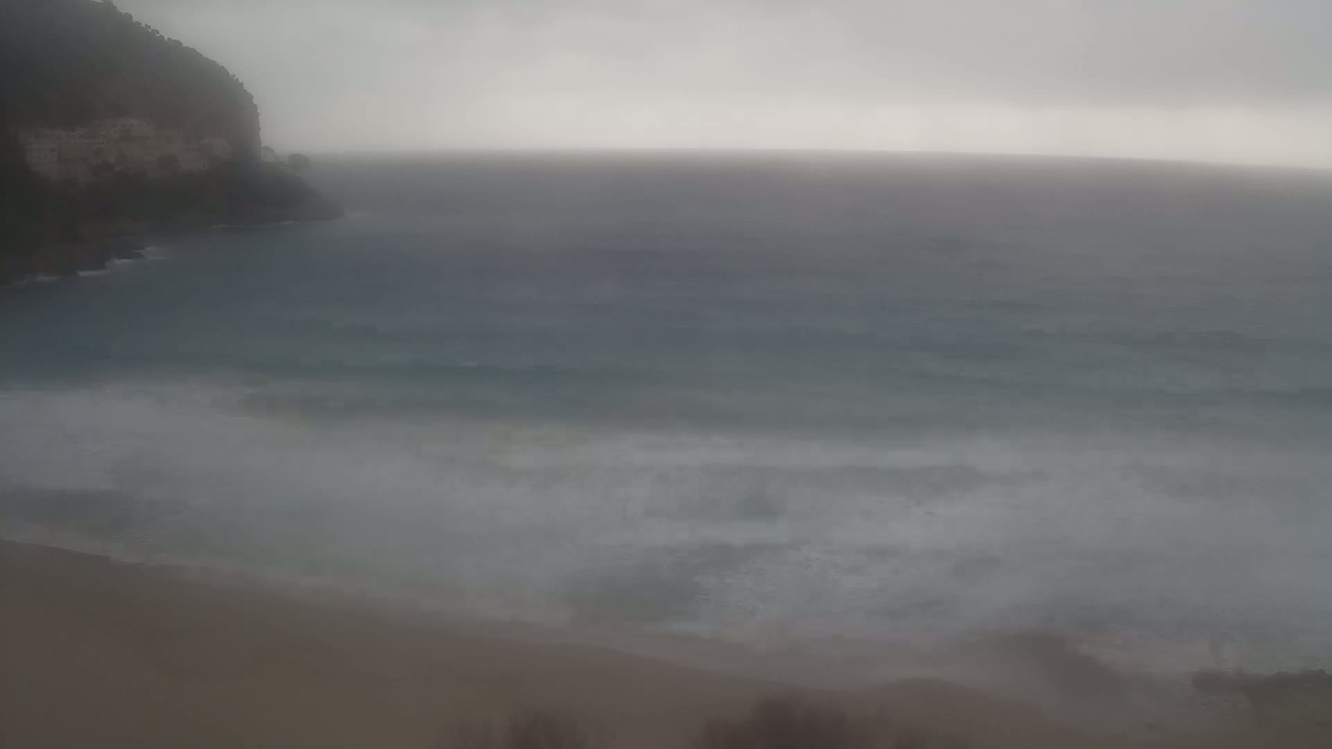 A hazy, overcast day shows a rocky cliff overlooking a choppy sea meeting a sandy beach.