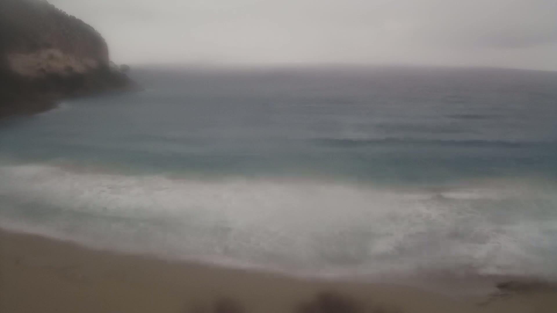 Waves crash on a sandy beach near a cliff face on an overcast day.