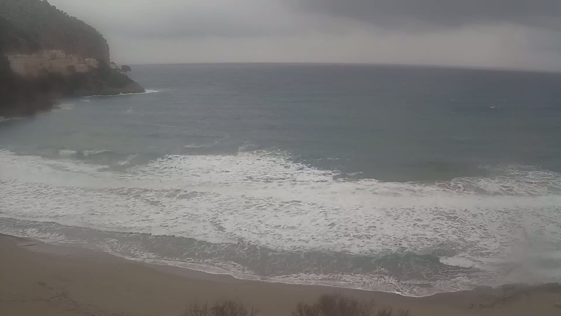 A sandy beach with white-capped waves rolls onto the shore under an overcast sky near a rocky cliff.