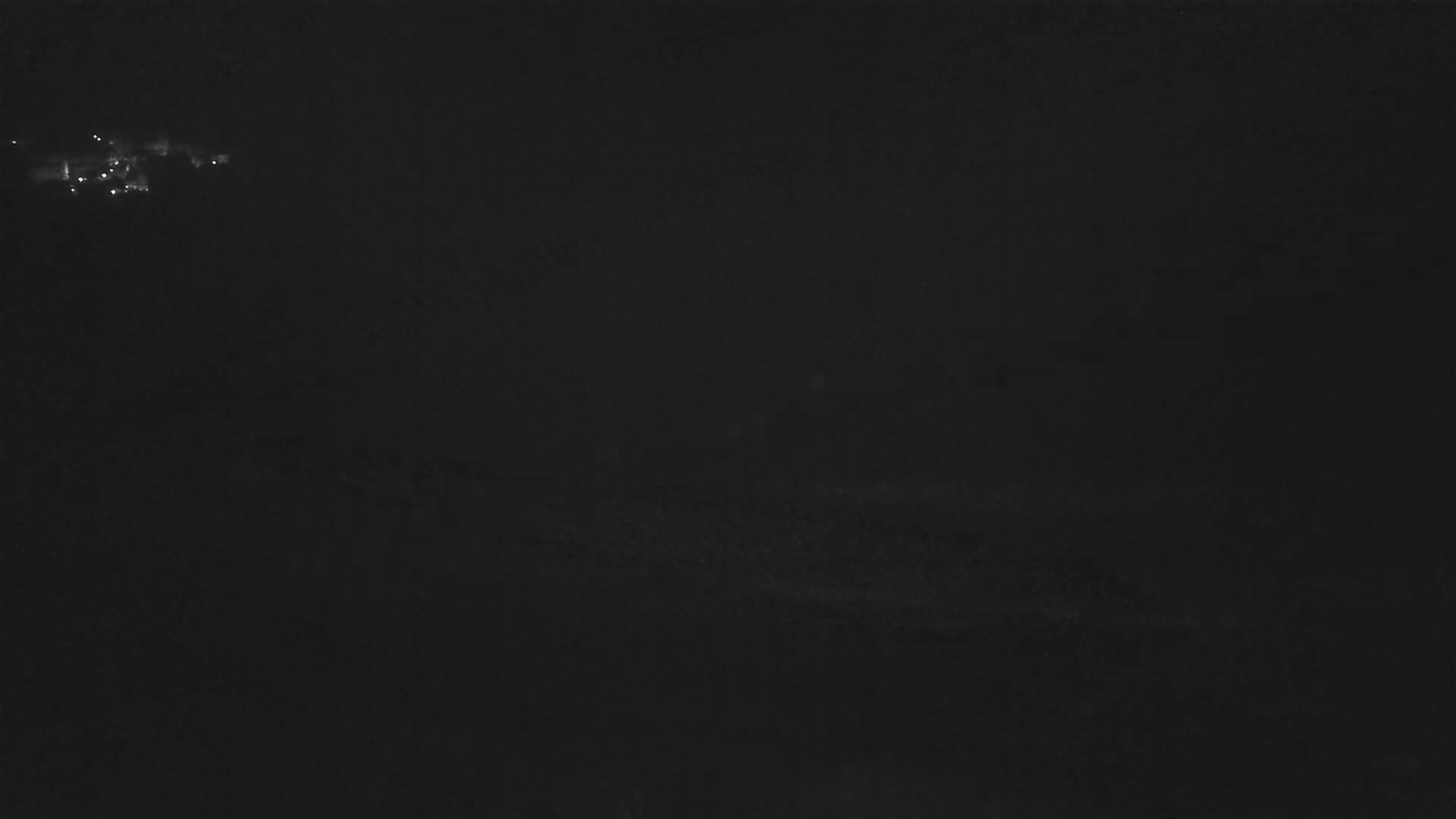 A dark, nighttime scene shows a distant cluster of faint lights on a hill, under a cloudy sky.