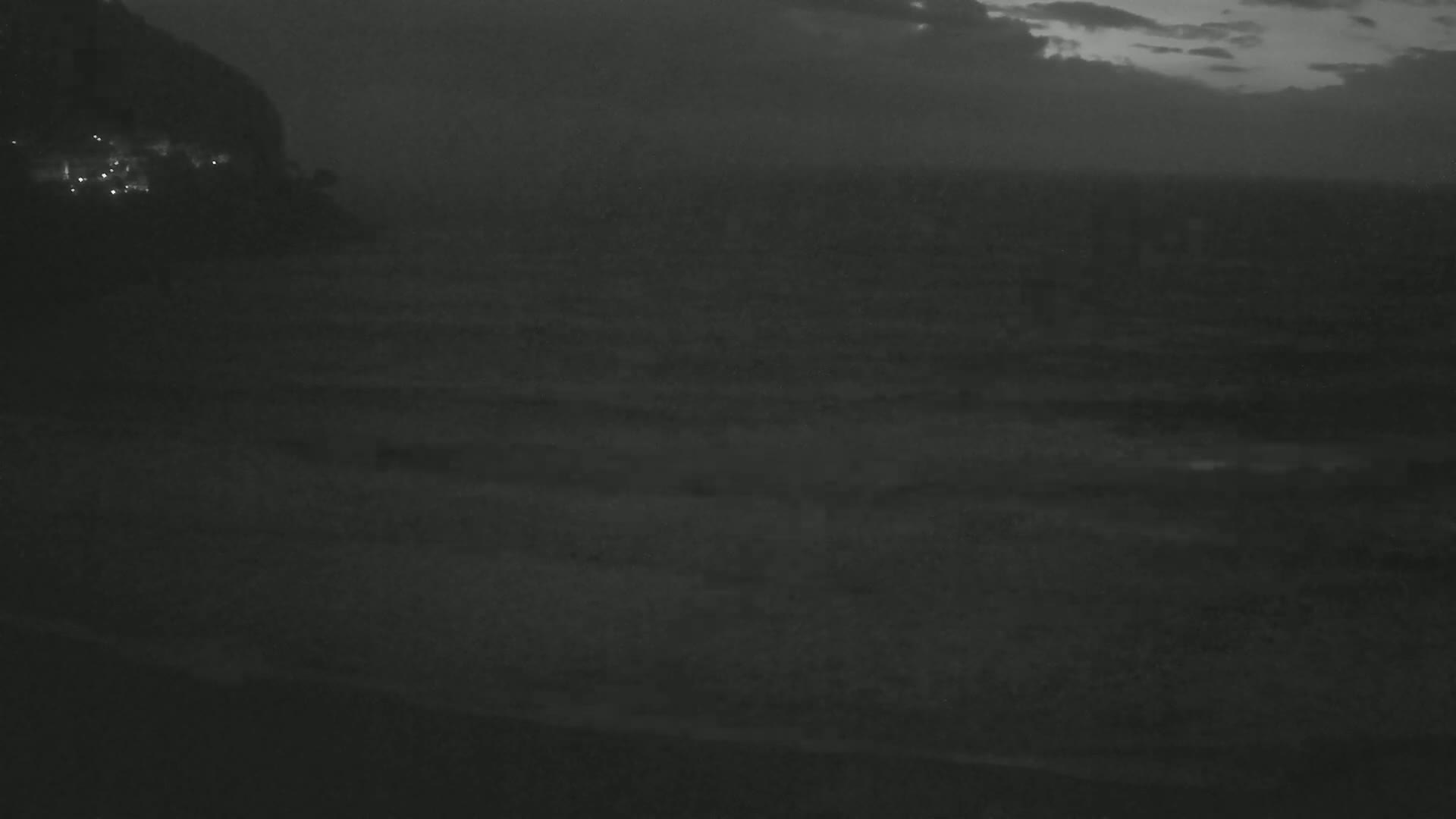 A dark, nighttime coastal scene shows a body of water with subtle waves under a cloudy sky.