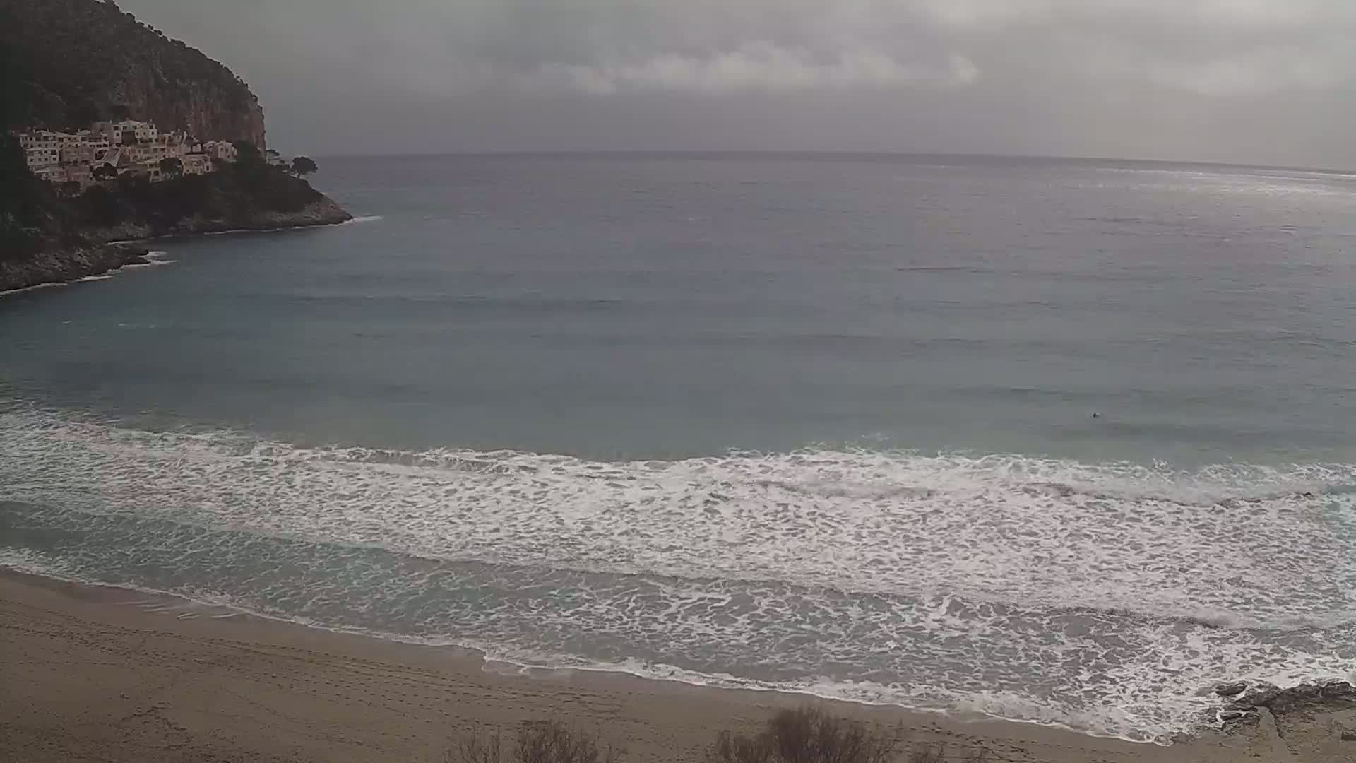 A sandy beach with small waves rolls onto shore under an overcast sky, near a rocky cliff with buildings on top.
