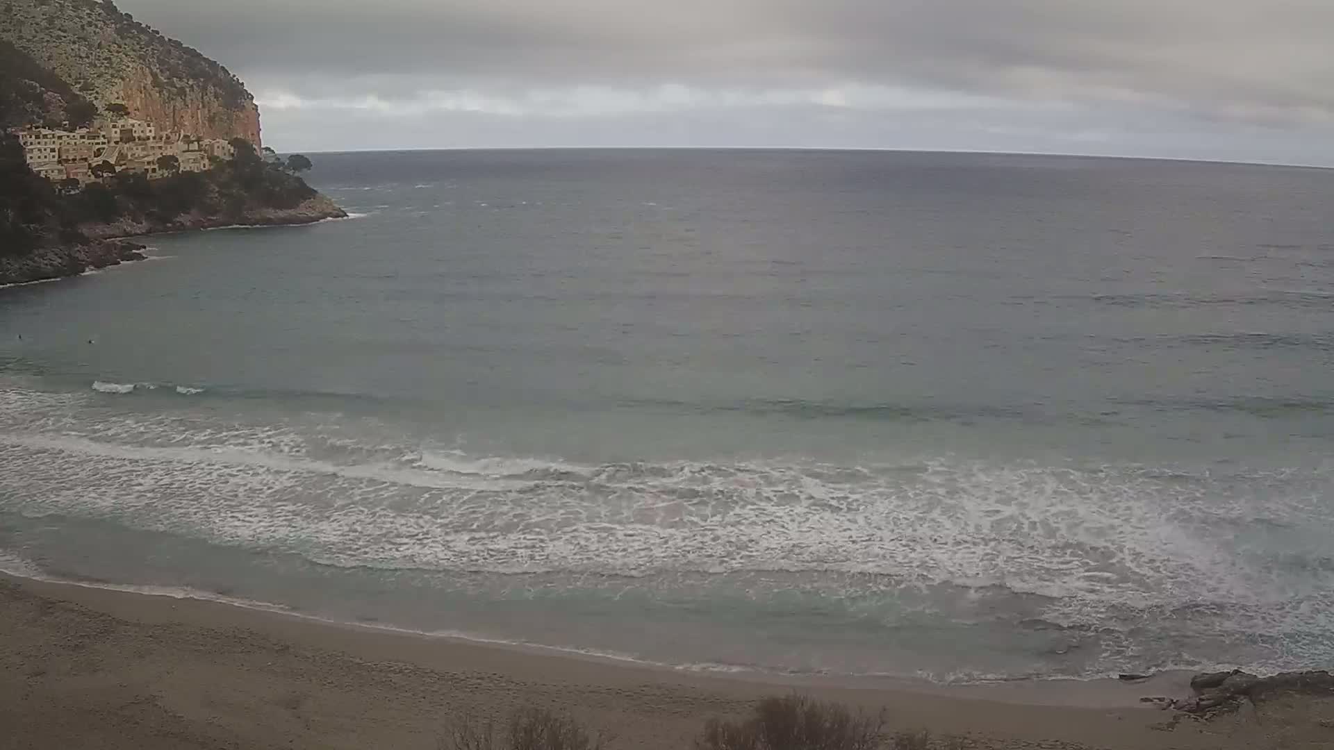 A sandy beach meets a calm, grey sea under an overcast sky, with a rocky cliff and buildings visible in the distance.