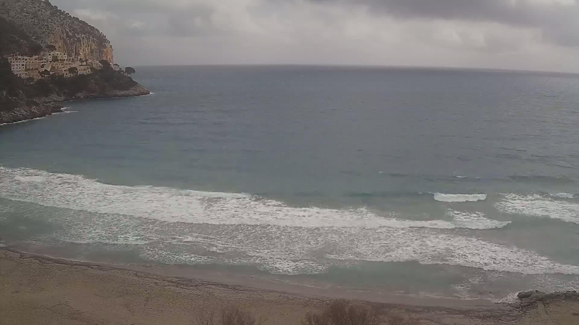 A sandy beach with small waves lapping the shore under an overcast sky, backed by a rocky cliff with buildings on top.