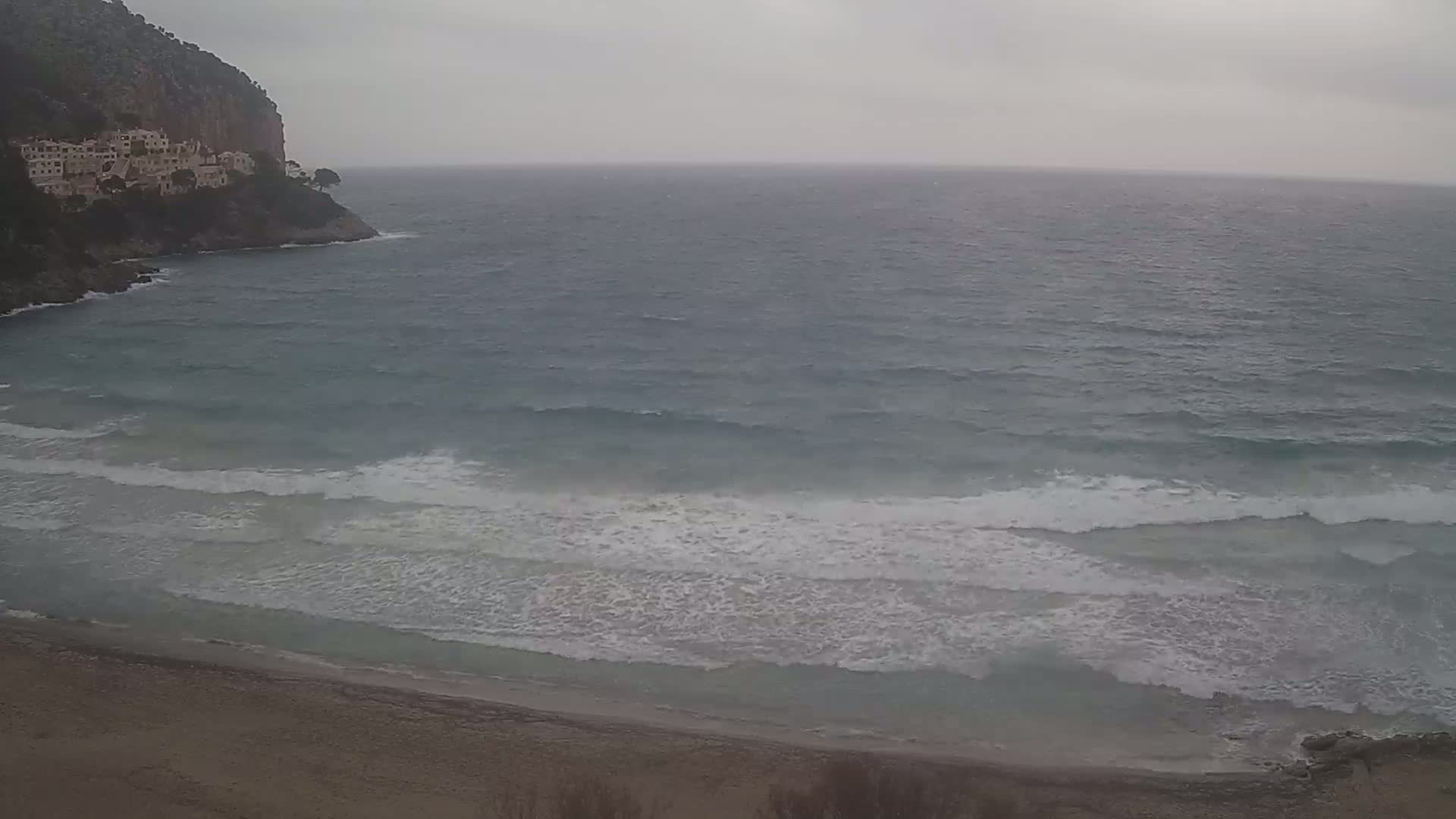 A sandy beach meets a choppy sea under an overcast sky, with buildings perched atop a cliff in the background.