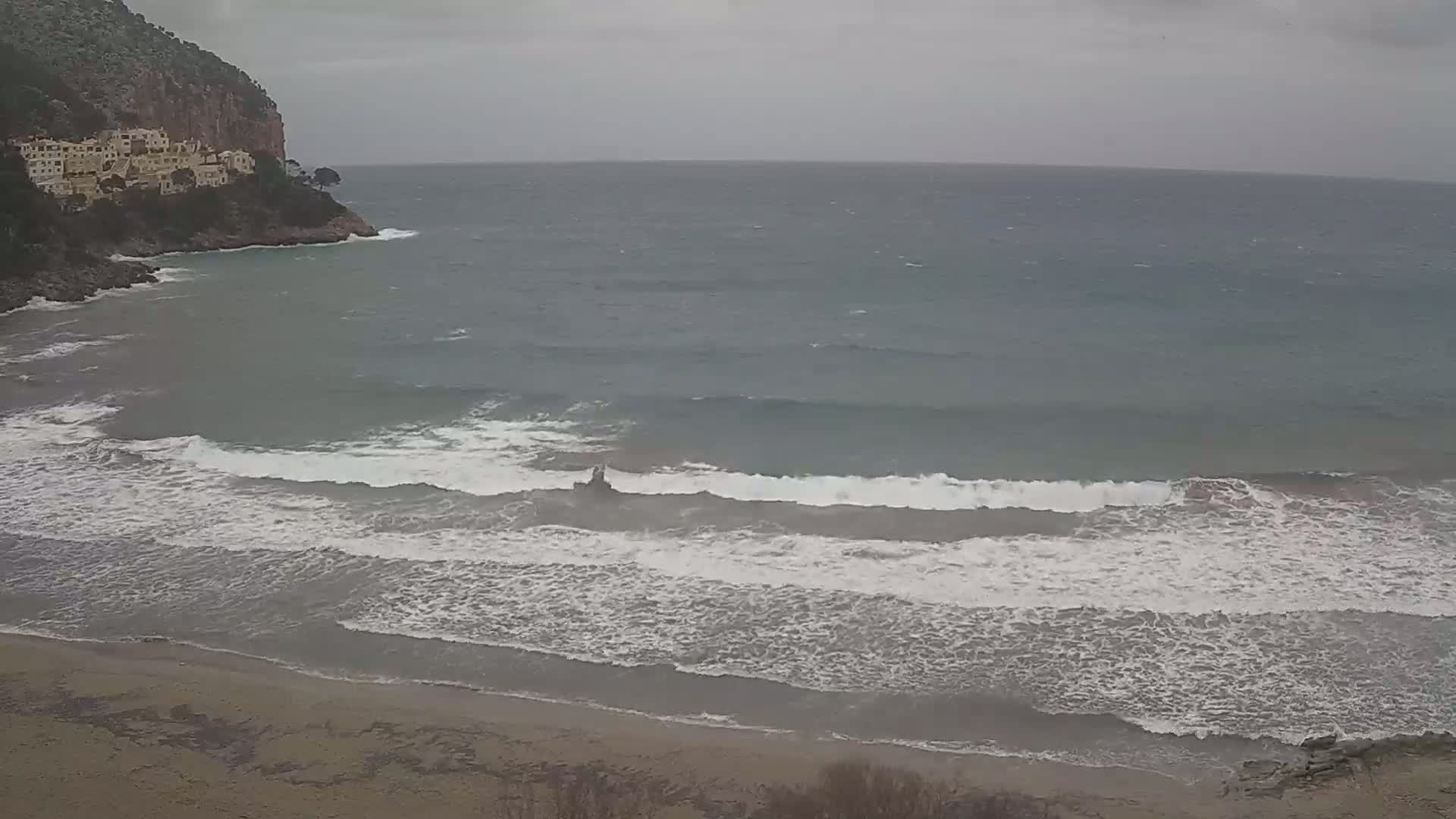 A sandy beach with moderate waves rolls onto the shore under an overcast sky, near a rocky cliff with buildings on top.