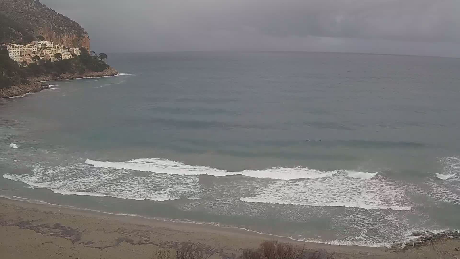 A sandy beach with small waves laps against the shore under an overcast sky, near a rocky cliff with buildings on top.