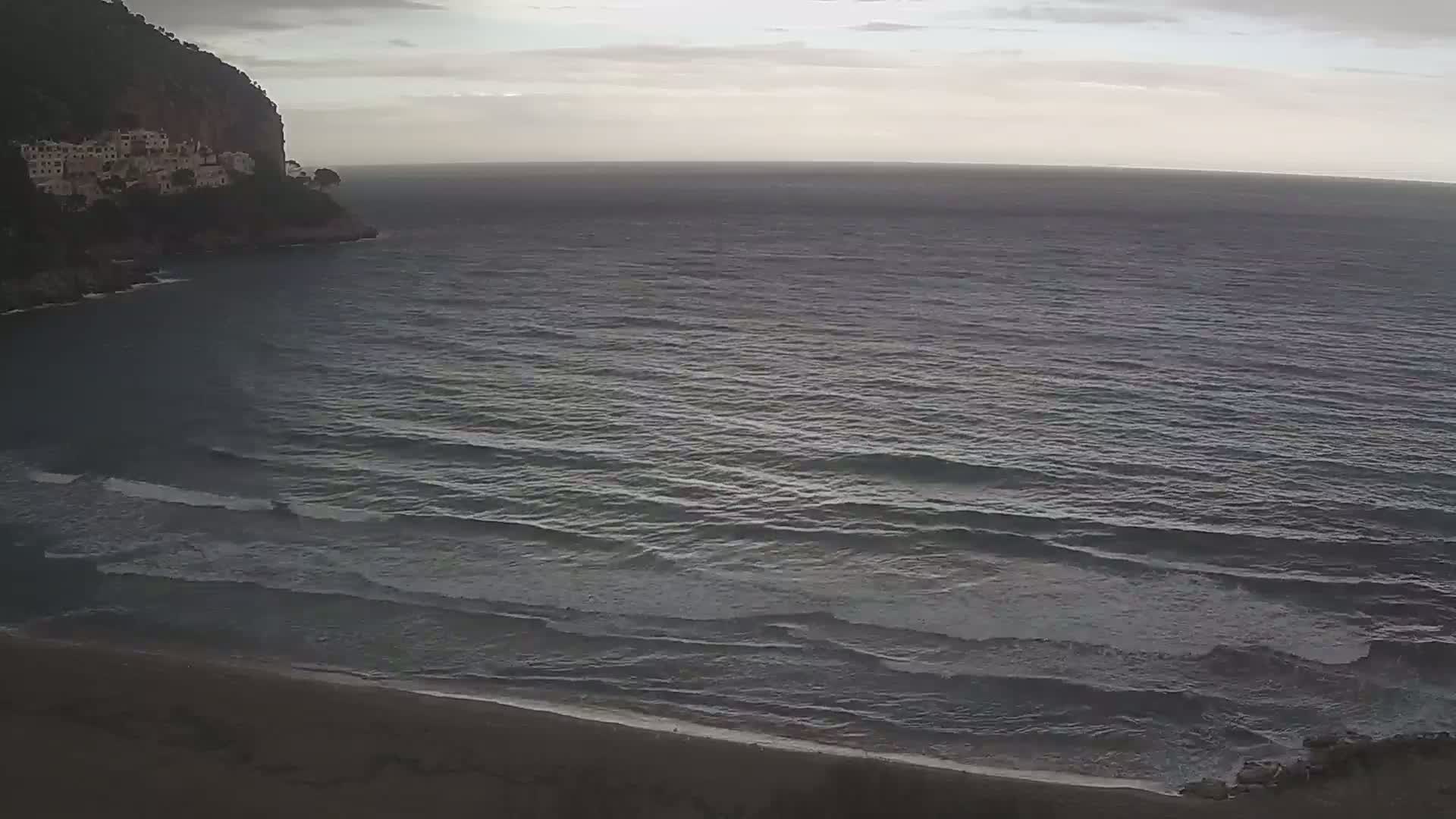 A dark, sandy beach meets a calm, grey sea under an overcast sky, with a cliff and buildings visible in the distance.