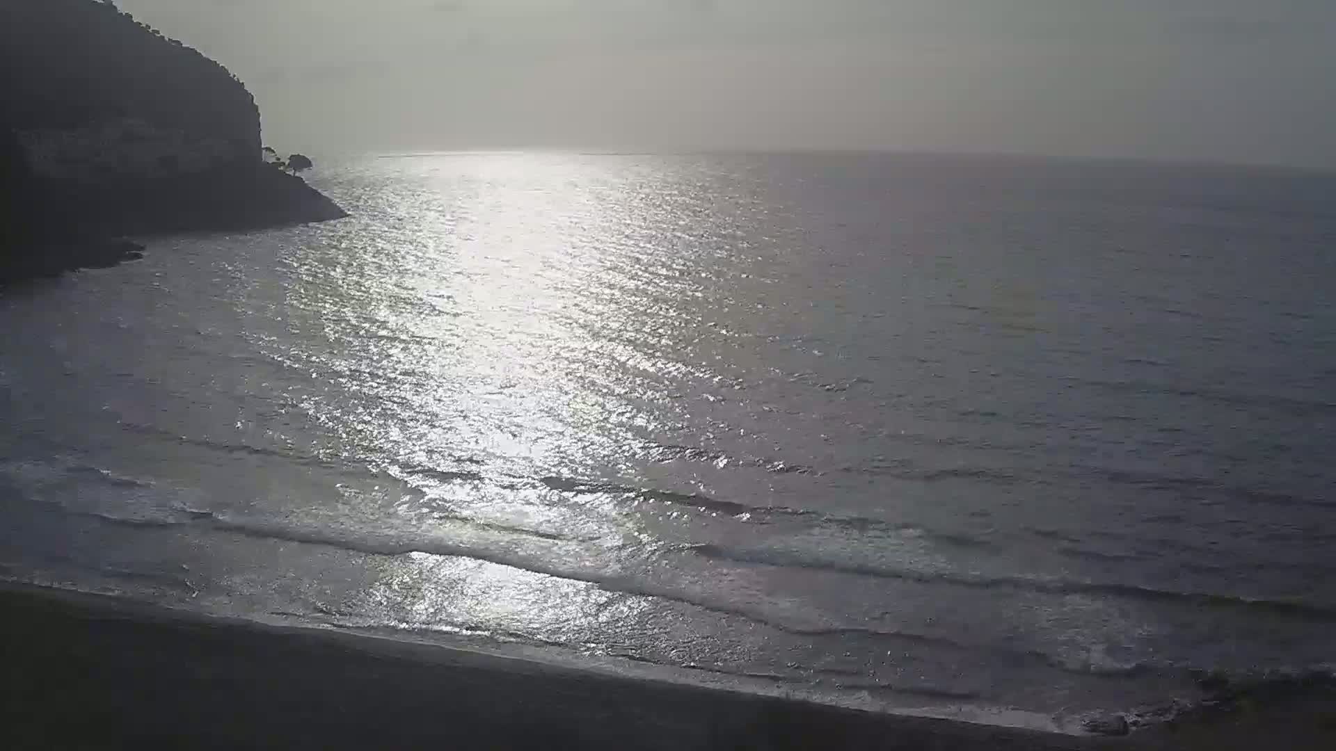 A dark, rocky cliff overlooks a calm sea with gentle waves and a bright reflection of sunlight on the water.