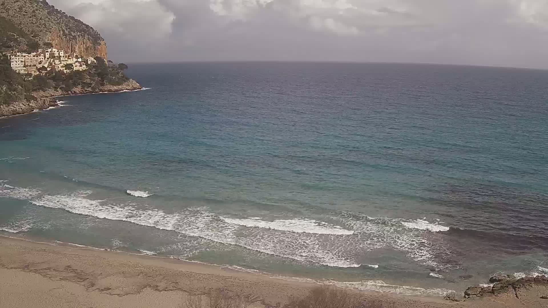 A sandy beach with gentle waves laps the shore near a rocky cliff, under a cloudy sky.