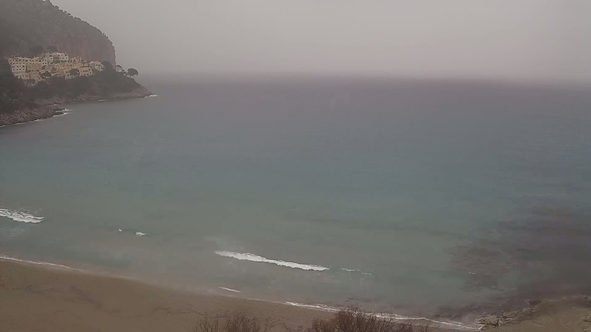 A hazy, overcast day shows a sandy beach meeting a calm, teal sea, which laps against a rocky shoreline with buildings perched on a cliffside.