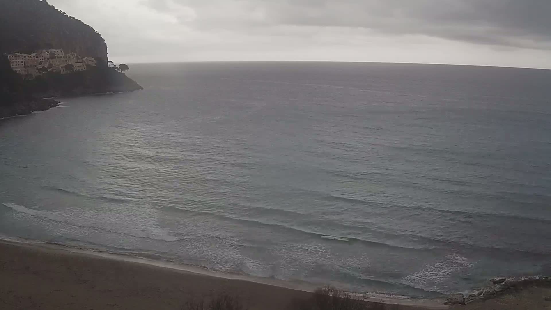 A sandy beach meets a calm, gray sea under an overcast sky, with a cliff and buildings visible on the left.