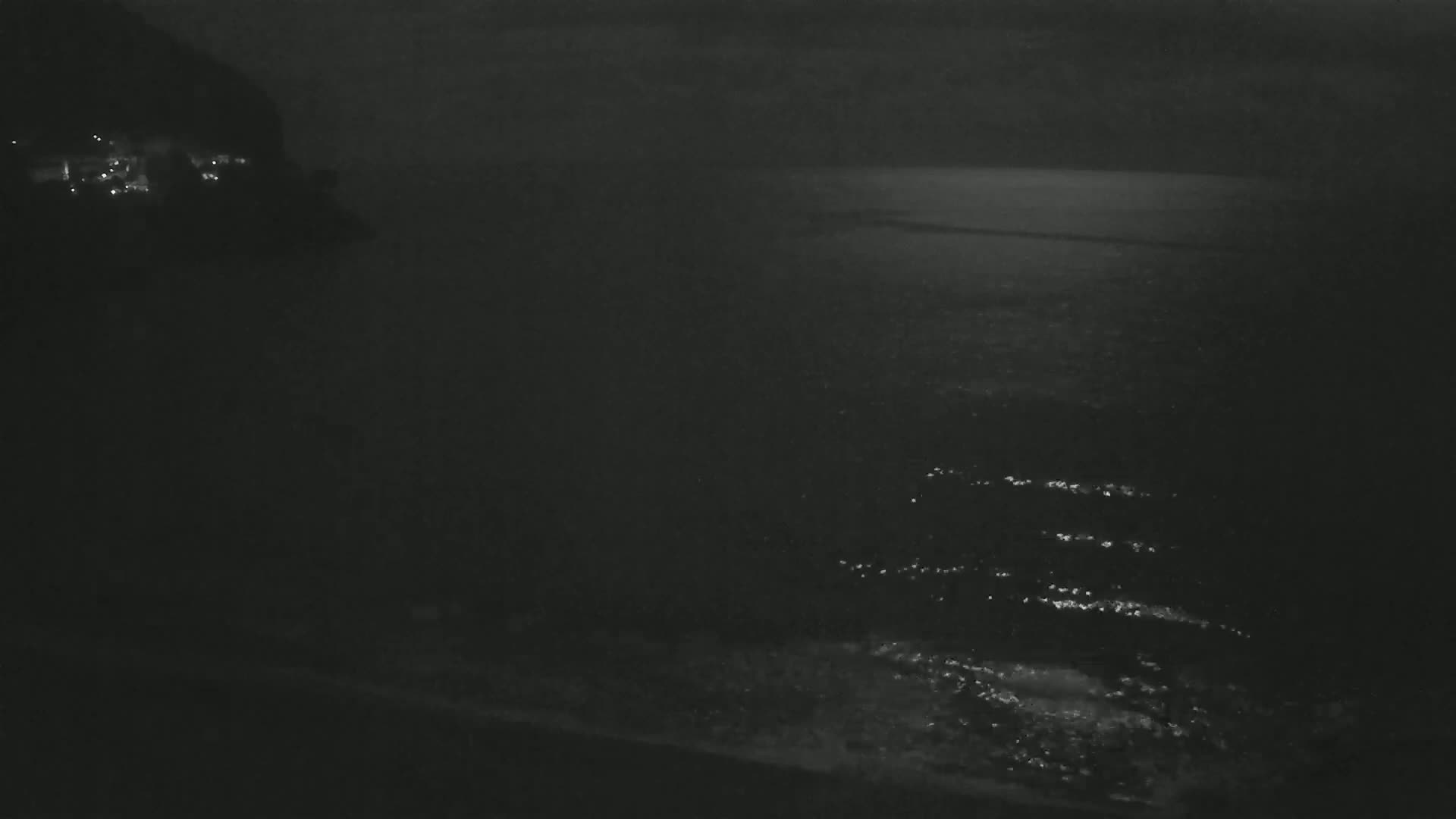 A dark, nighttime coastal scene shows a body of water with gentle waves lapping against a shore, with a dimly lit landmass visible in the background.