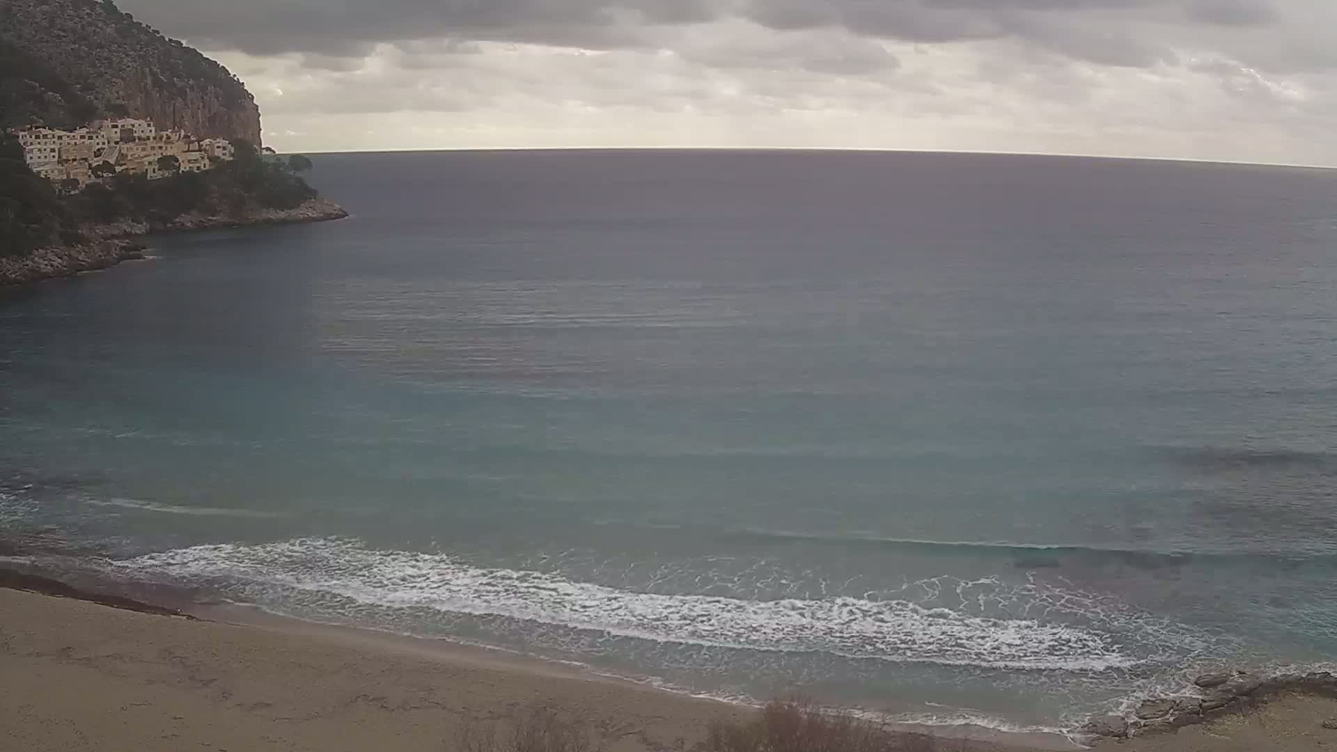 A sandy beach meets a calm, teal sea under a cloudy sky, with a cliff and buildings visible in the distance.