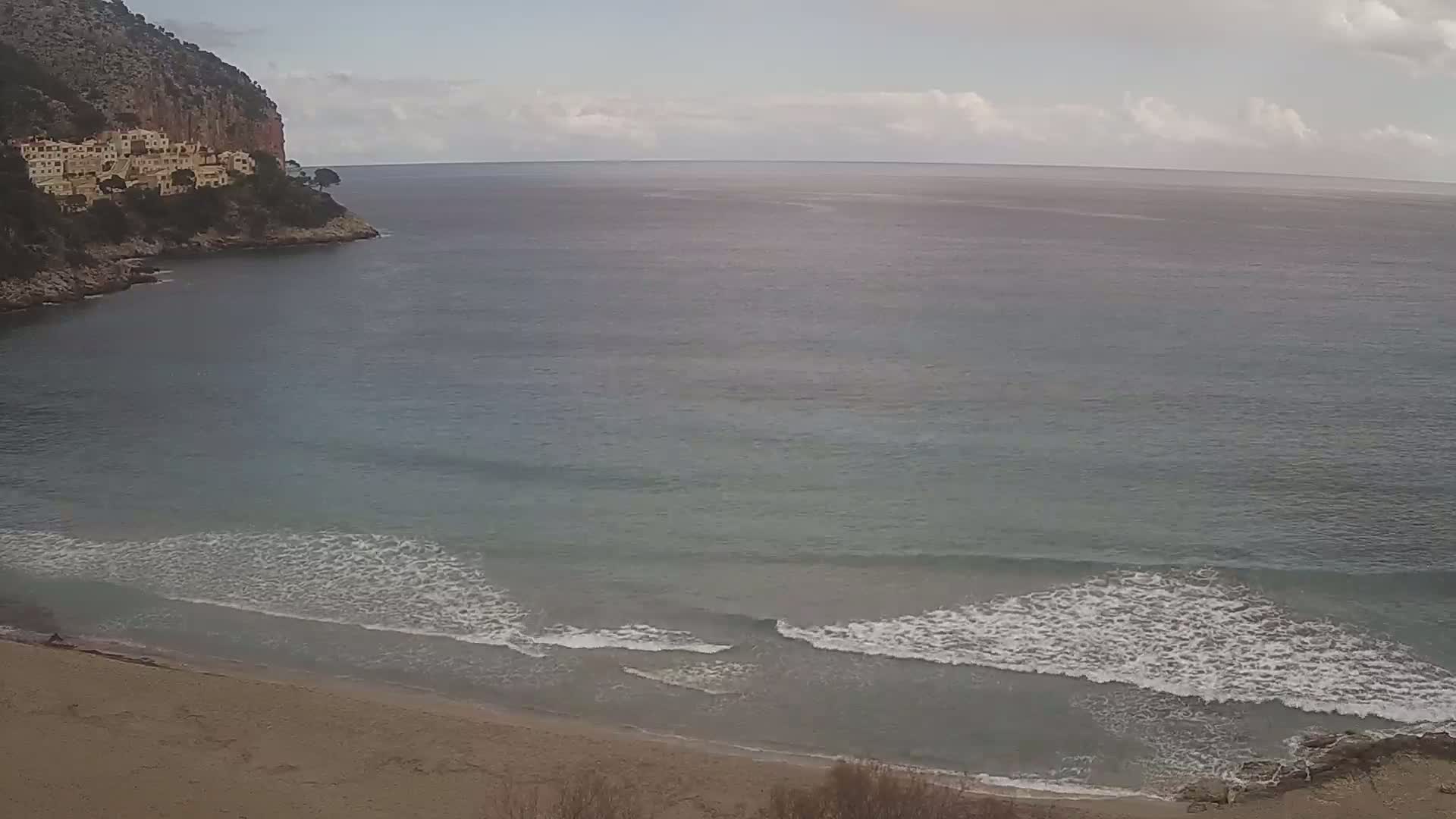A sandy beach with small waves laps against the shore, under a mostly cloudy sky, near a rocky cliff with buildings on top.