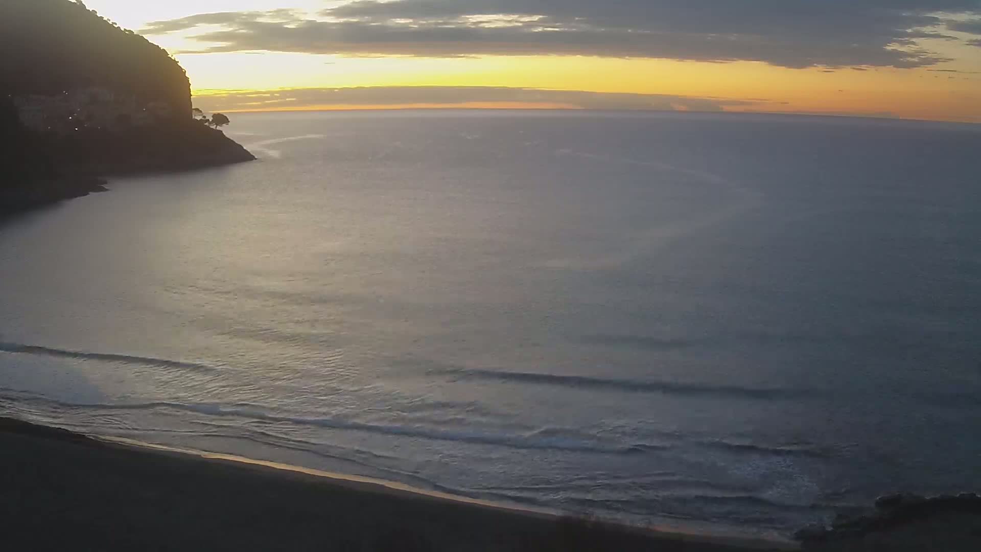 A dark, rocky cliff overlooks a calm ocean with small waves gently lapping a sandy beach at sunset.