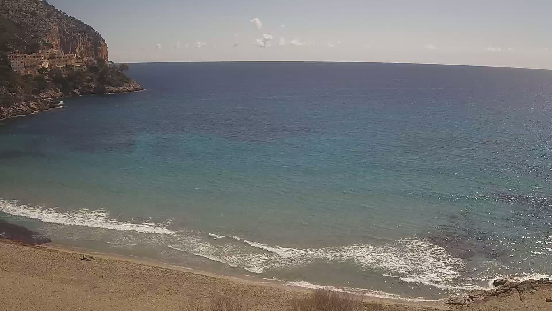 A sandy beach with gentle waves laps against the shore under a mostly clear sky, next to a calm turquoise sea and a rocky cliff with buildings on top.
