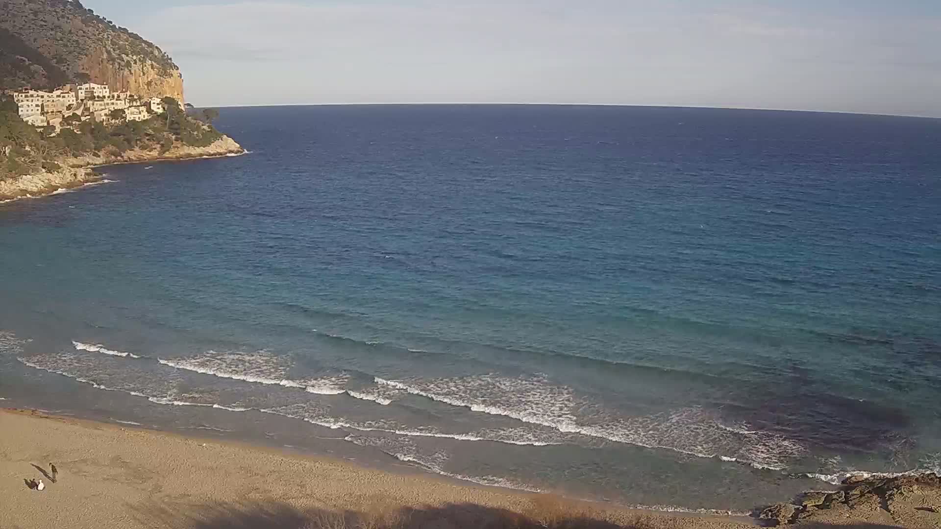 A sandy beach with gentle waves laps the shore, meeting a calm sea under a clear sky; buildings cling to a rocky cliffside in the background.