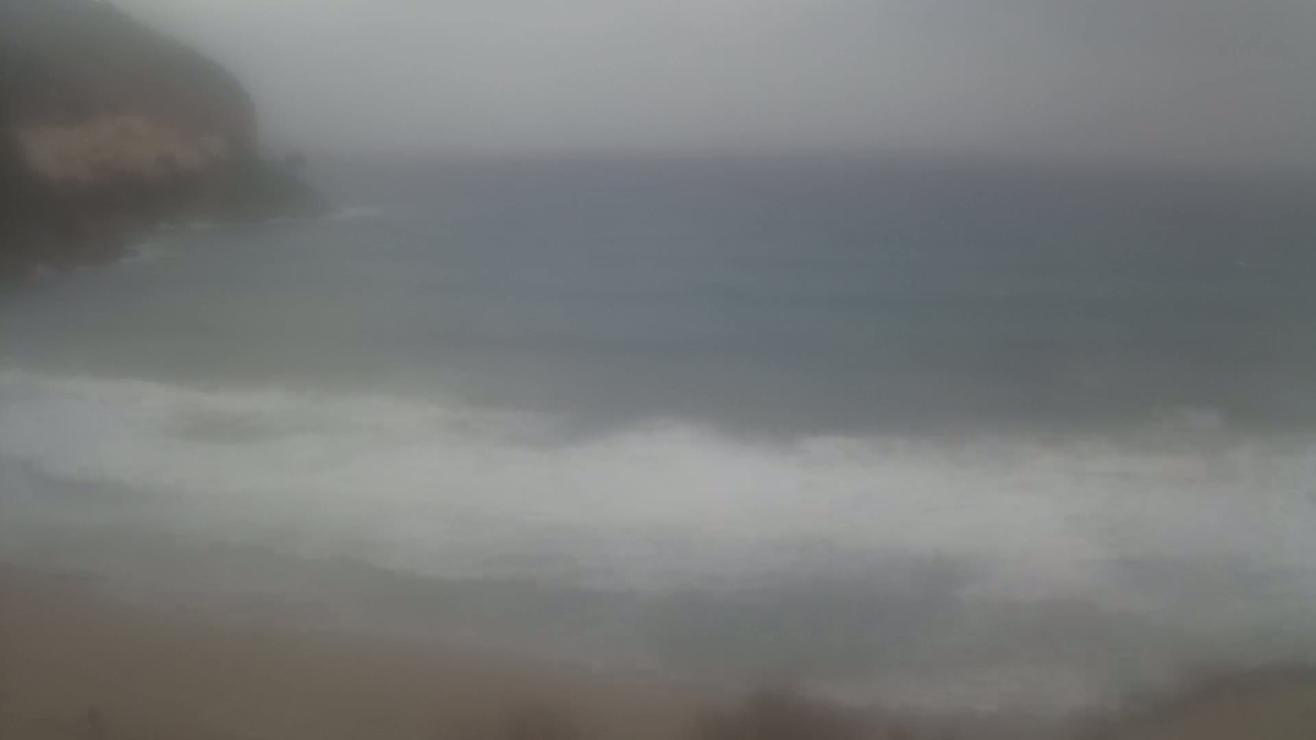 A hazy, overcast sky overlooks a beach with waves breaking on the shore near a dark cliff.