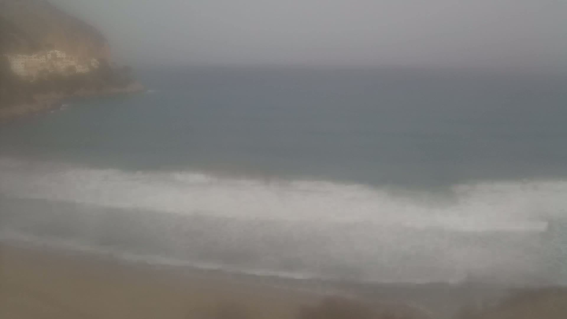 A hazy, overcast day shows a sandy beach with waves breaking on the shore near a rocky cliff.