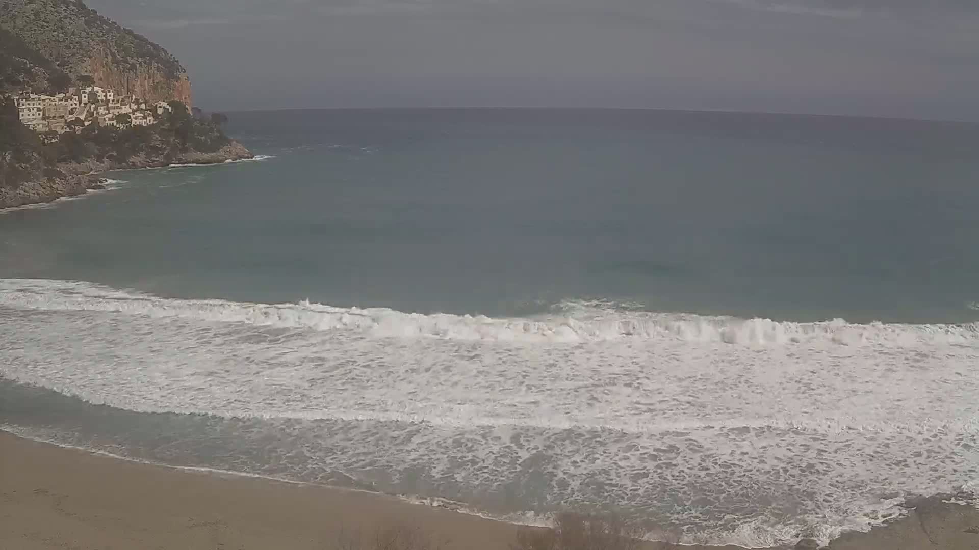 White-capped waves crash on a sandy beach near a rocky cliff with buildings on top, under an overcast sky.