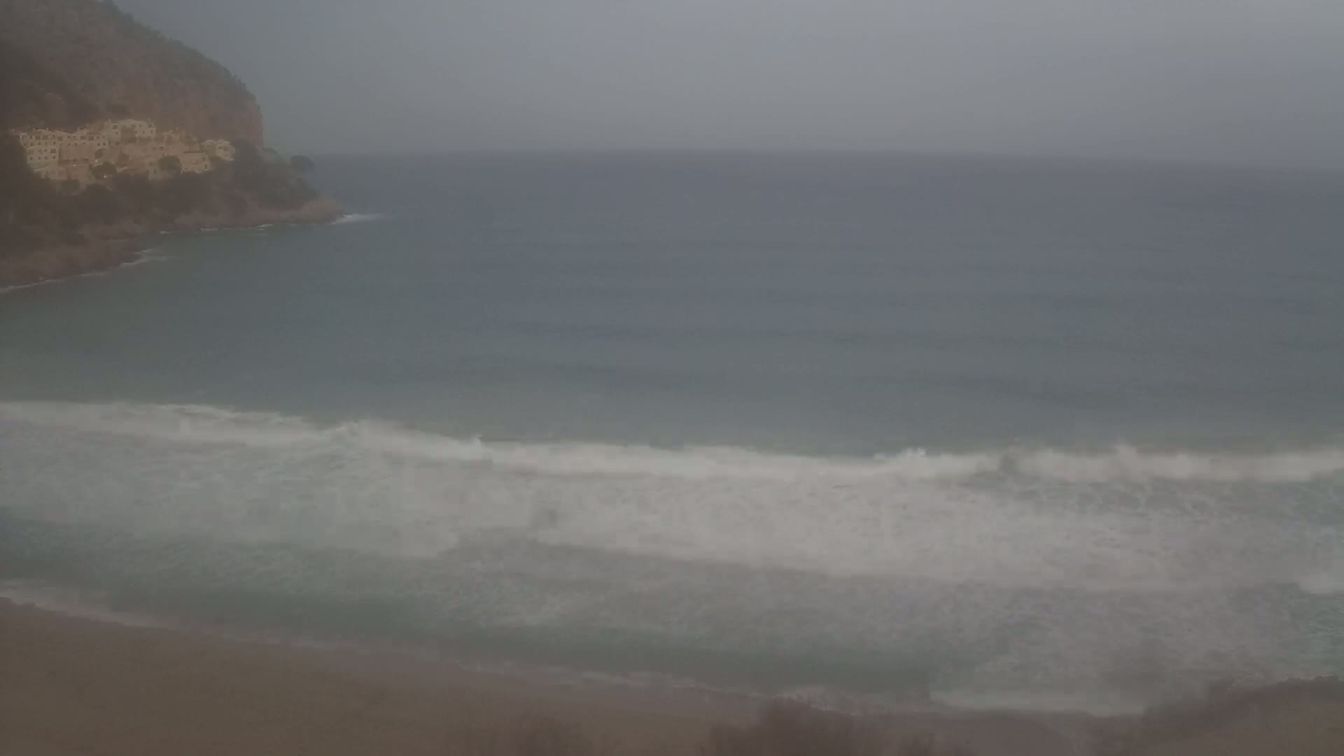 A hazy, overcast day reveals a sandy beach with waves breaking on the shore near a rocky cliff with buildings.