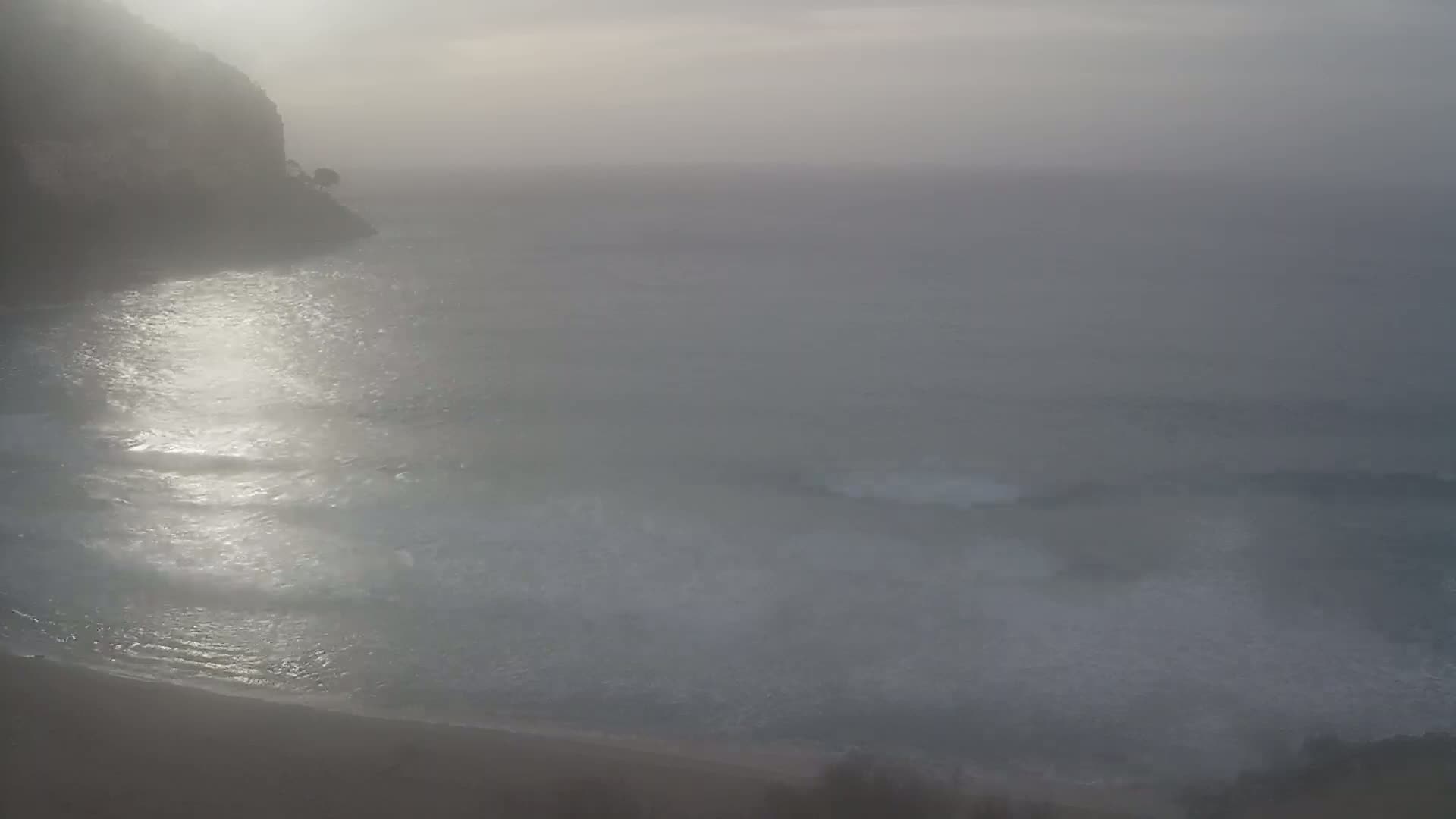 A foggy coastal scene shows a dark cliff, a beach, and a gray sea with gentle waves.