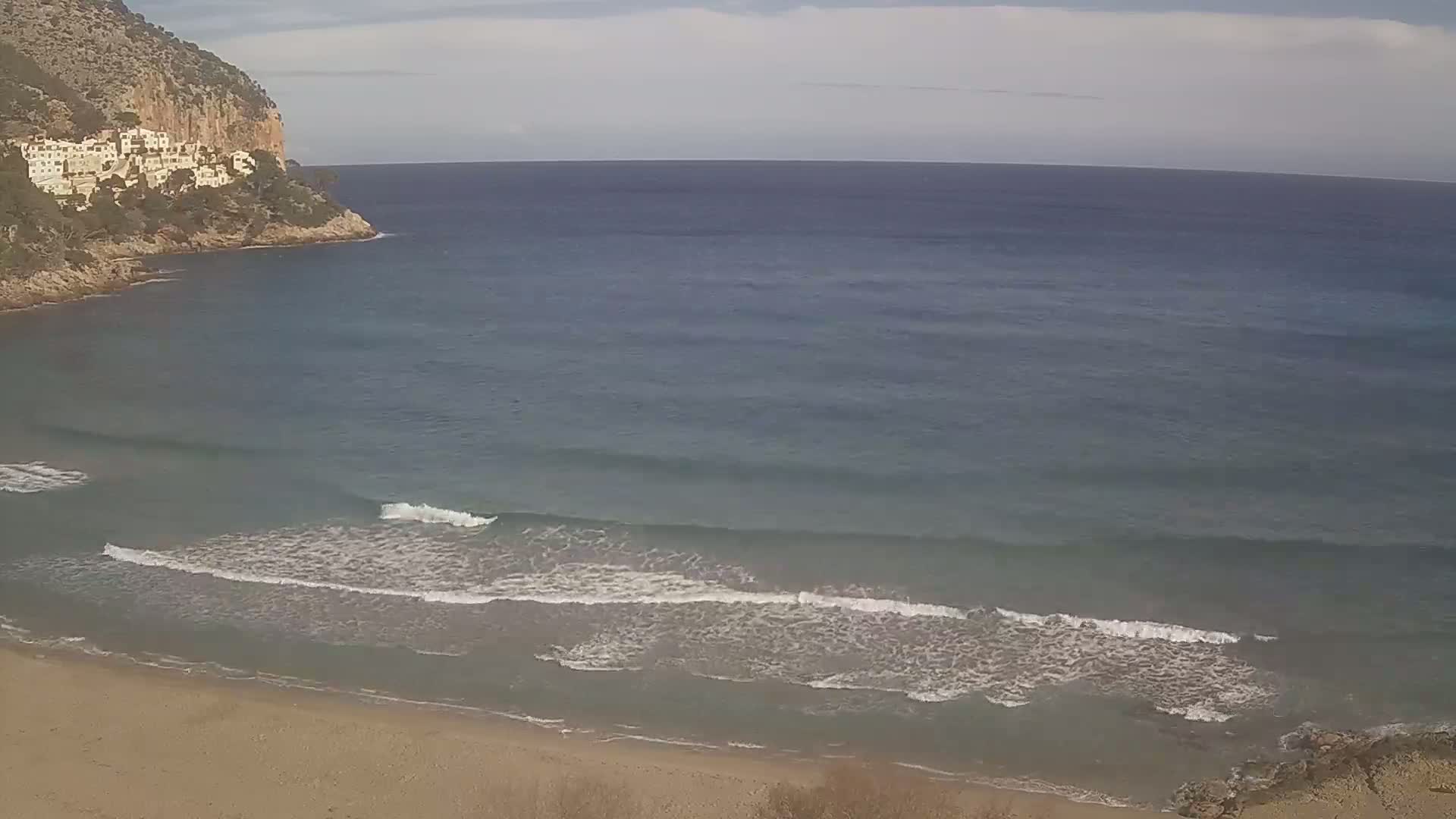 A sandy beach with small waves gently lapping the shore, backed by a rocky cliff with white buildings, under a partly cloudy sky.