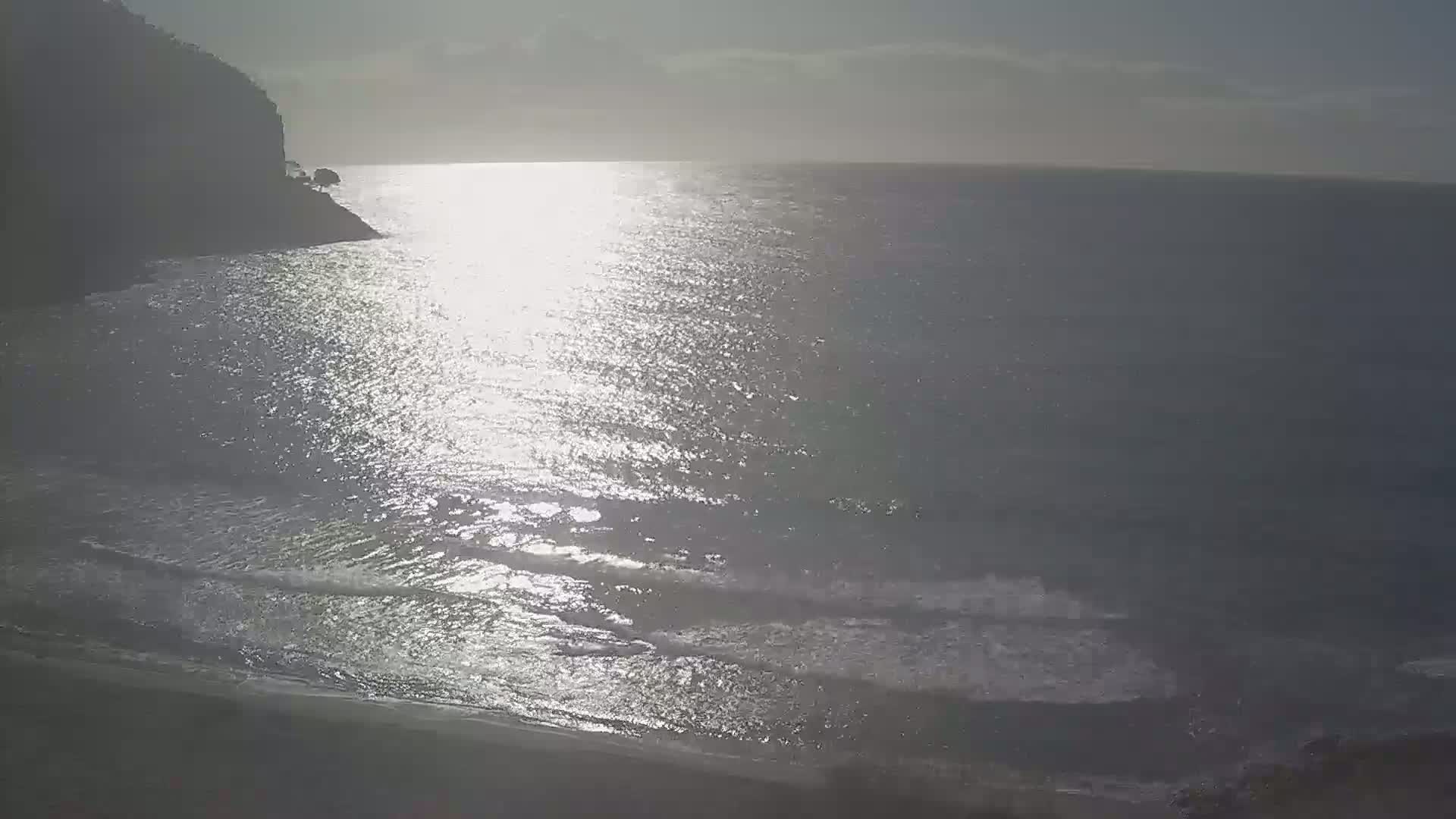 A bright, sunlit ocean with small waves laps a sandy shore near a dark cliff.