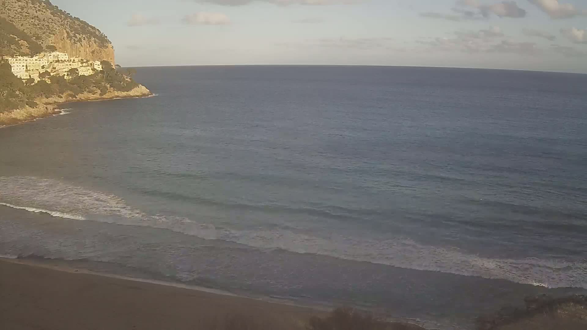 A sandy beach meets a calm, gray-blue sea under a partly cloudy sky, with a cliff featuring buildings visible in the distance.