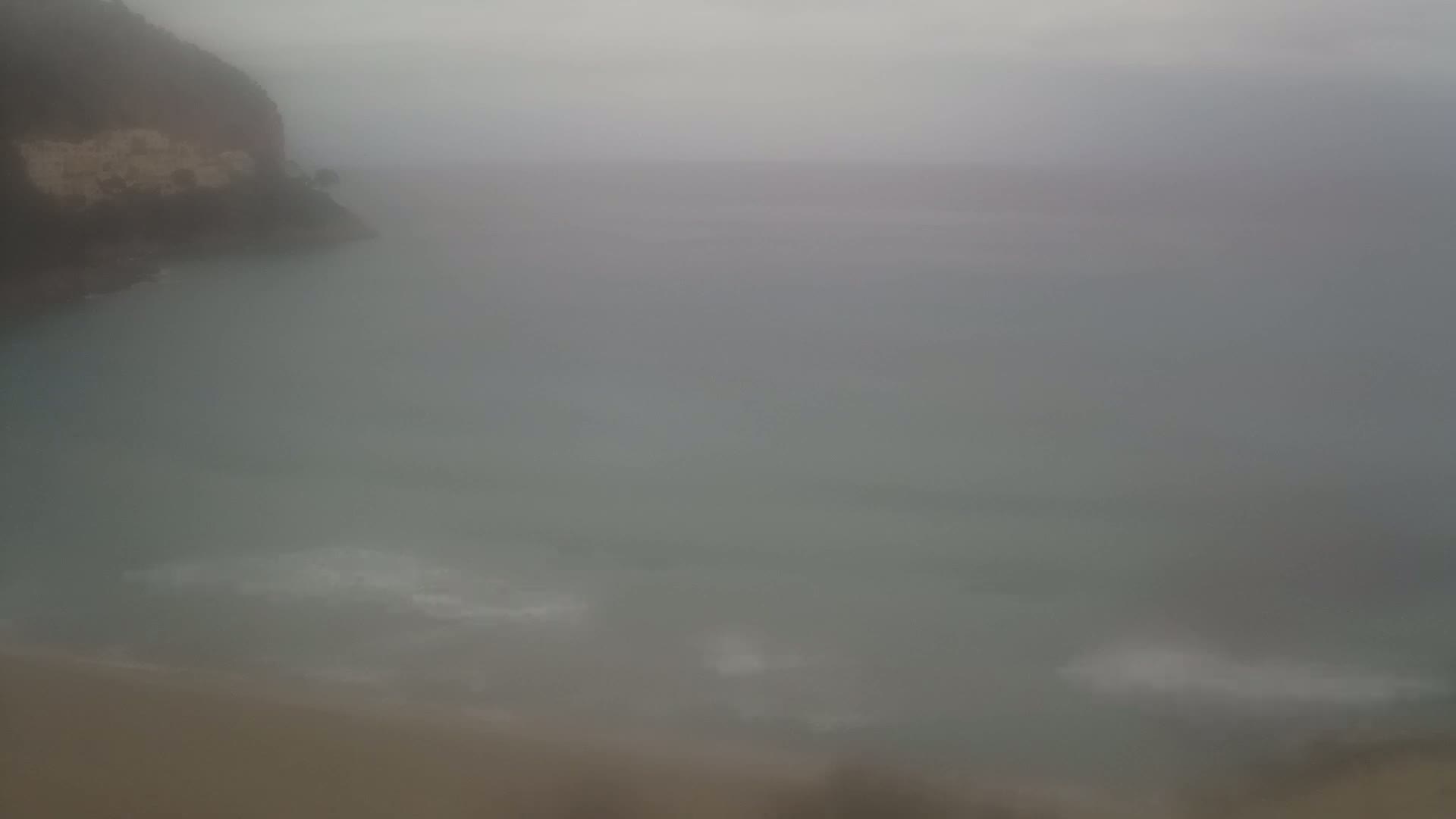 A hazy, overcast sky hangs over a calm body of water that meets a rocky, cliff-like shoreline.