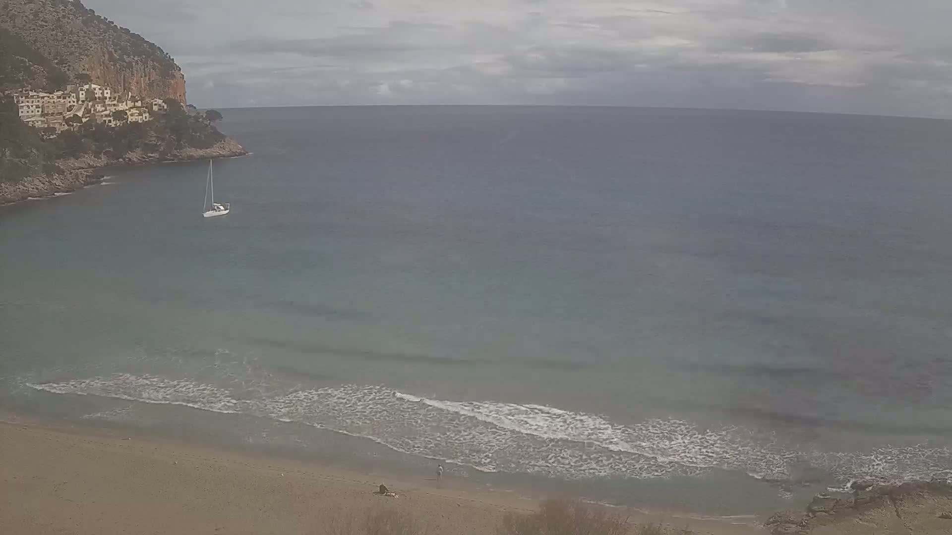 A sandy beach with small waves meets a calm bay, where a sailboat is anchored near a rocky cliff with buildings on top, under an overcast sky.