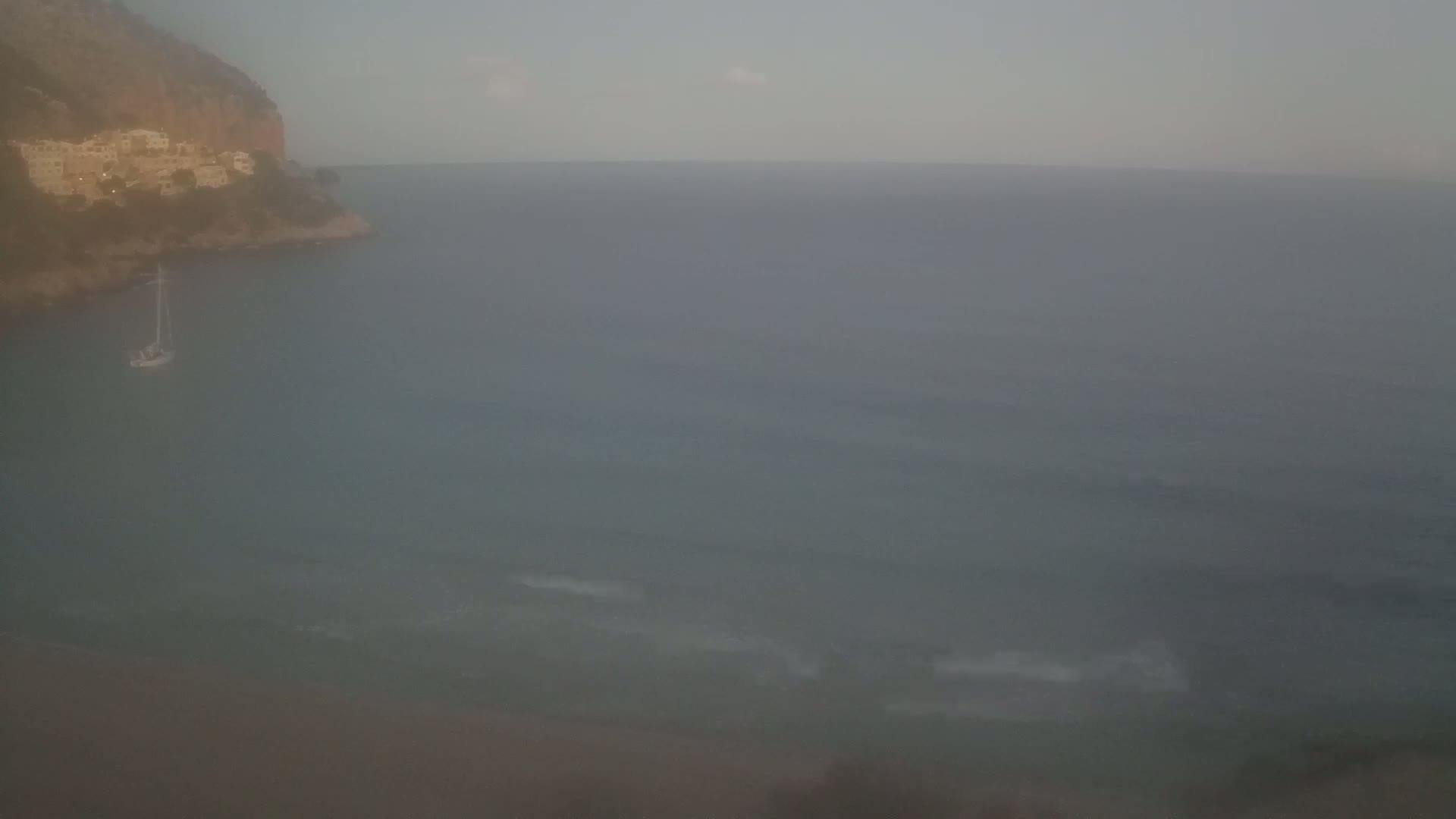 A hazy view shows a sailboat anchored near a rocky shoreline with buildings on a hillside, overlooking a calm sea.
