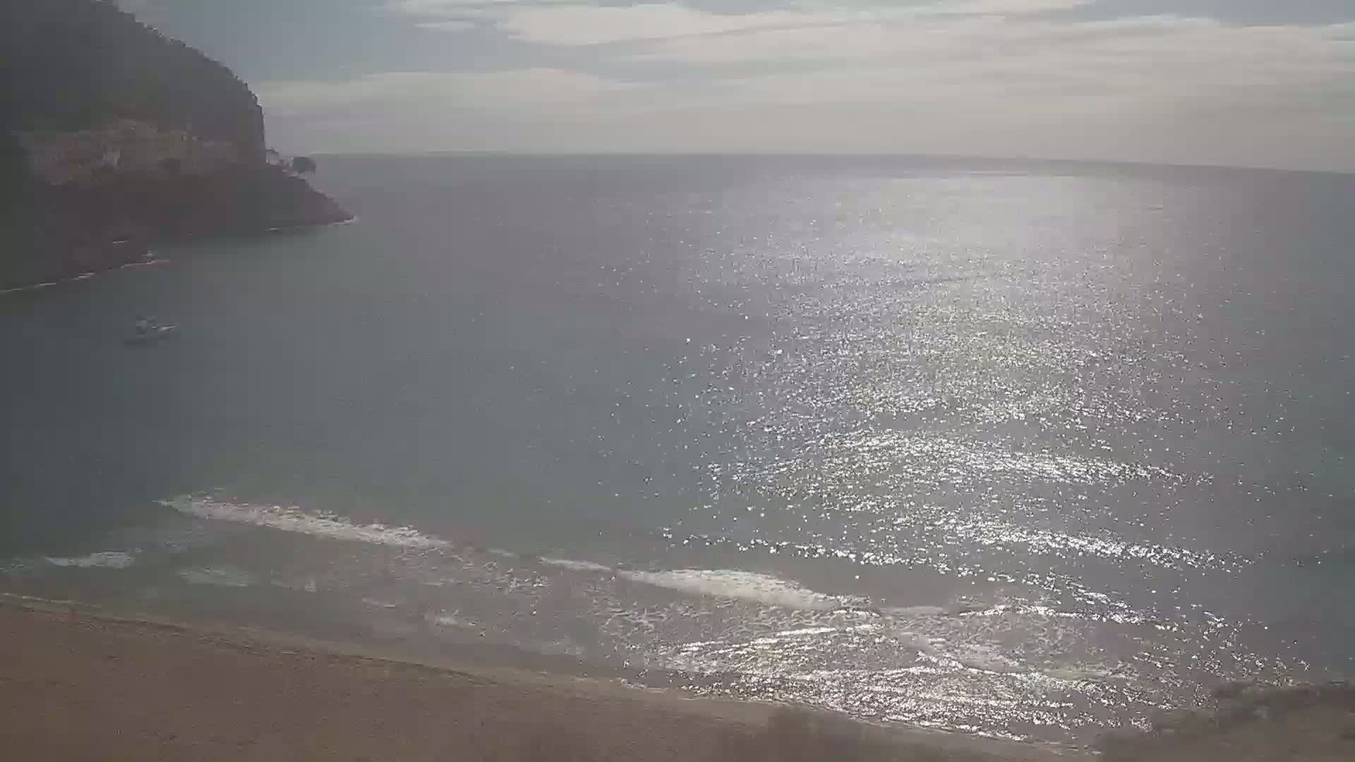 A sandy beach meets a calm, sunlit sea near a dark cliffside.