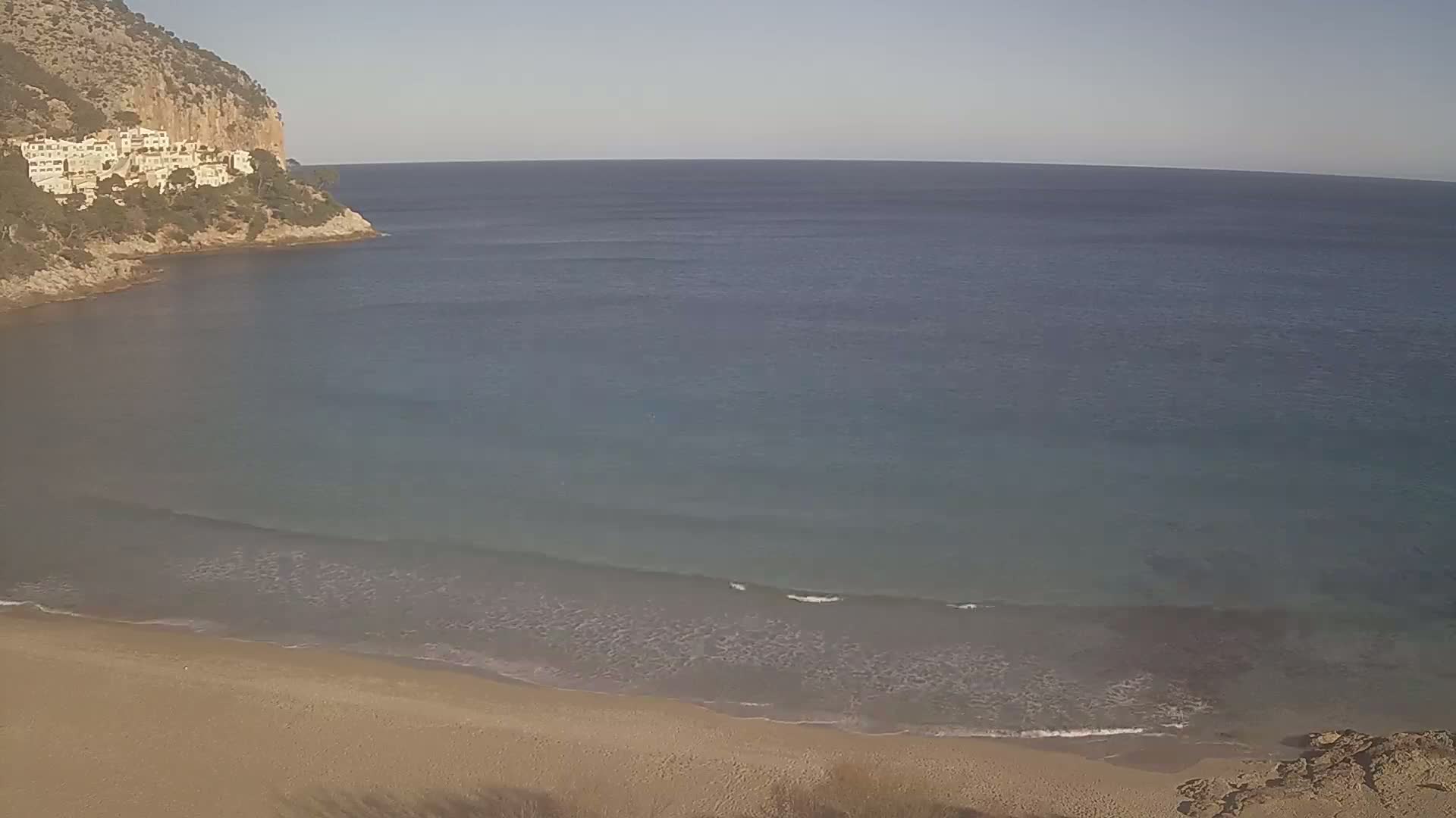 A sandy beach meets a calm, clear ocean under a bright, sunny sky, with a rocky cliff and buildings visible in the background.