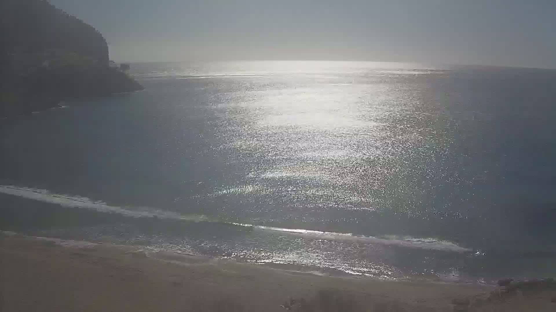 A sandy beach meets a calm, sunlit ocean next to a dark, rocky cliff on a hazy day.