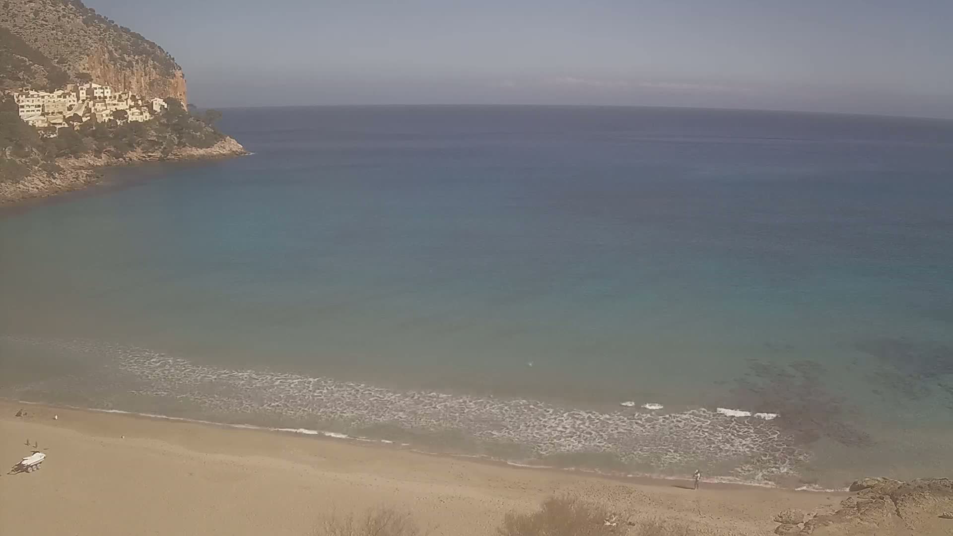 A sandy beach with gentle waves laps a calm, turquoise sea, backed by a rocky cliff with buildings on it under a clear, sunny sky.
