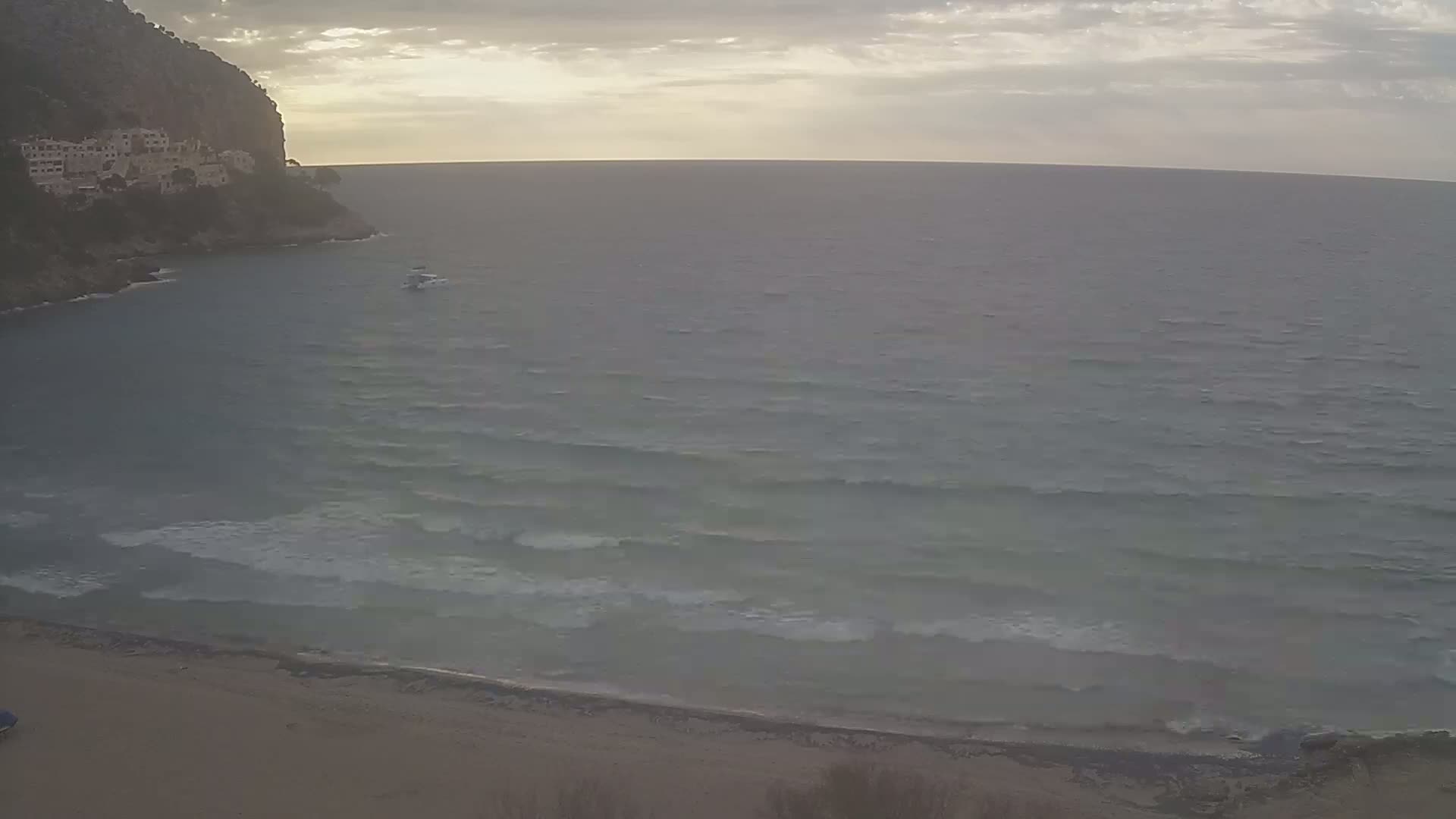 A sandy beach meets a calm sea with small waves under a cloudy sky, with a small boat visible on the water and buildings on a cliff in the background.