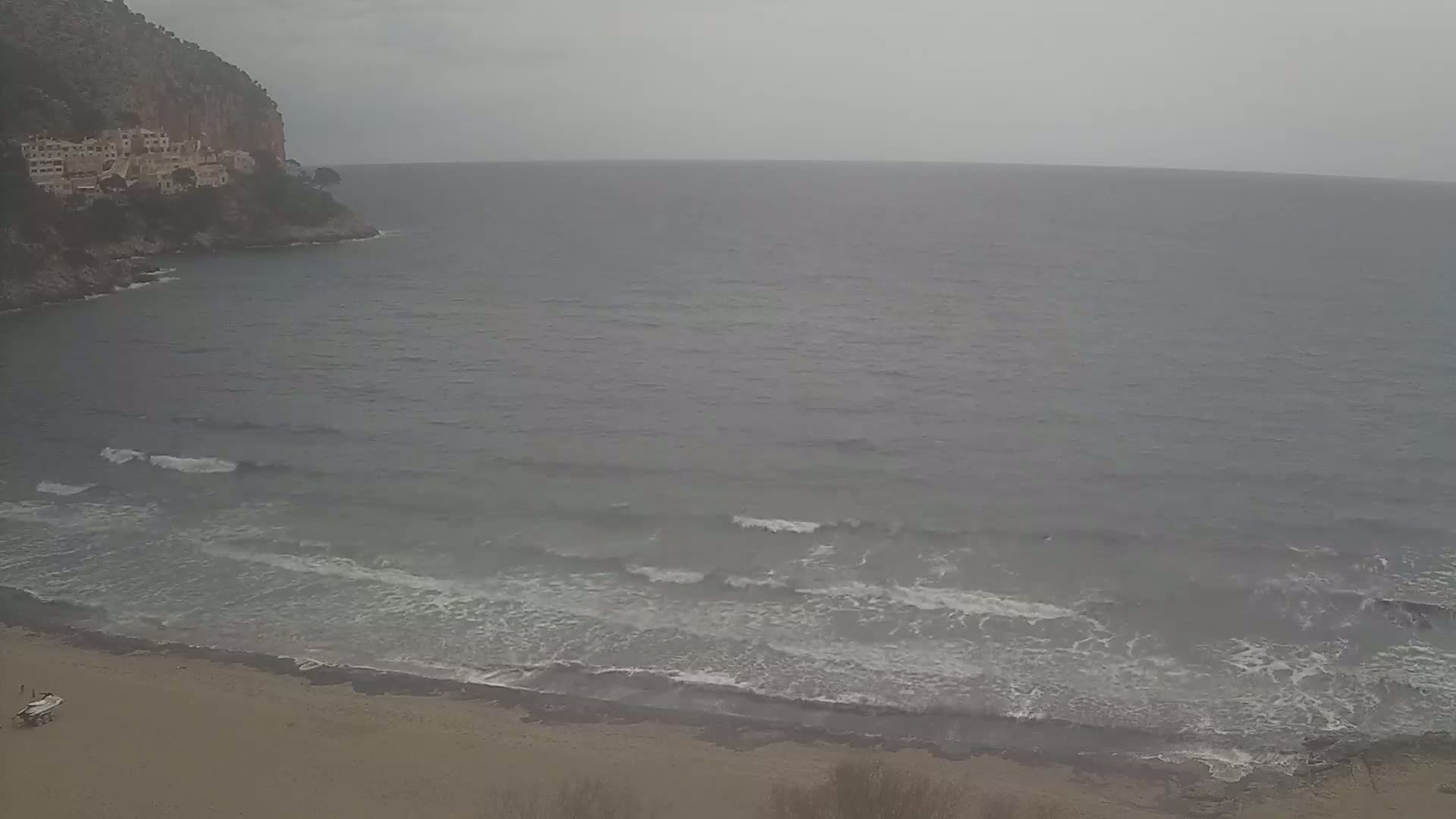 A sandy beach with small waves rolls onto the shore below a cliff with buildings under an overcast sky.