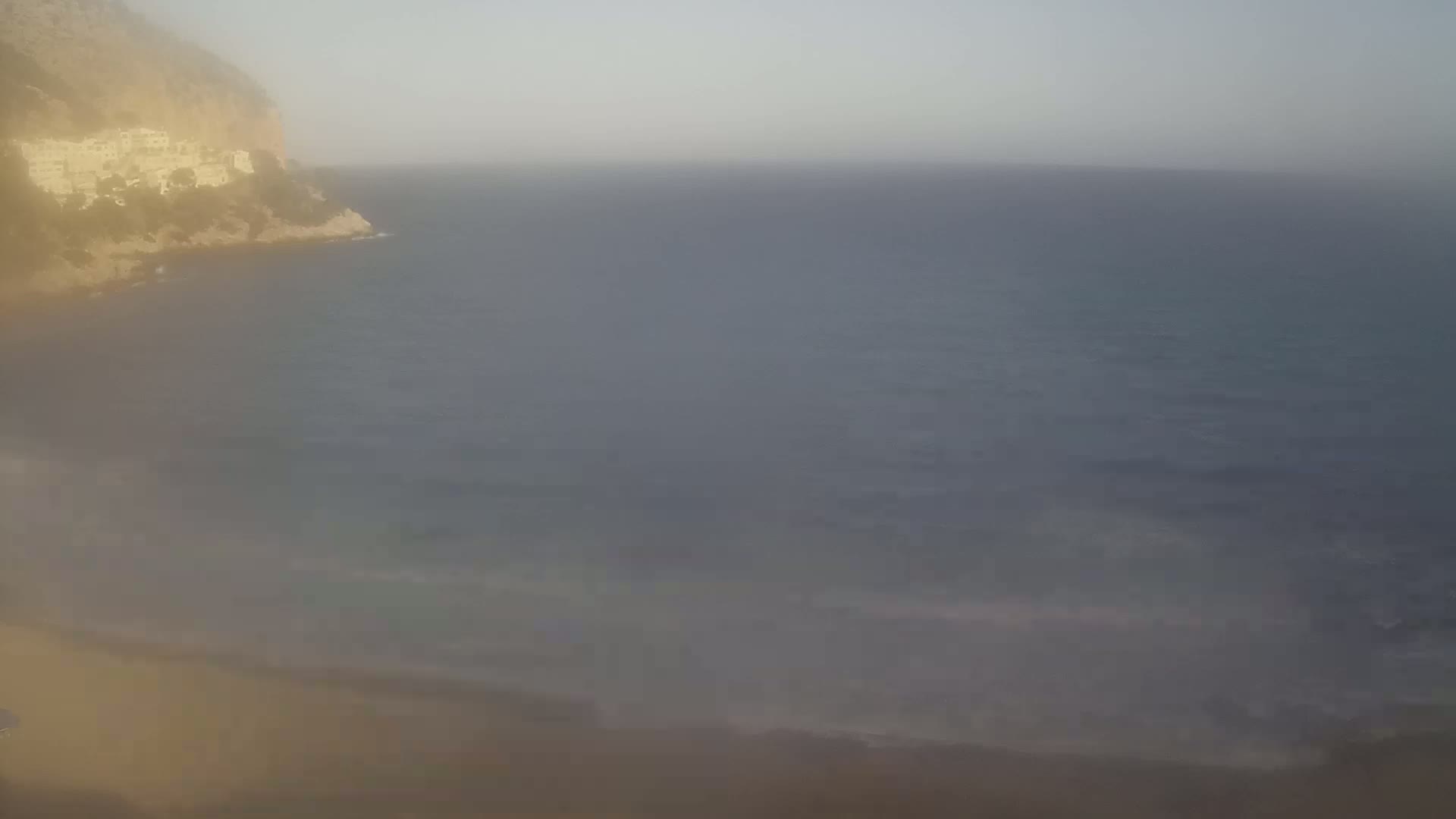 A hazy, slightly overcast day shows a sandy beach meeting a calm sea, with a cliff and buildings visible in the distance.