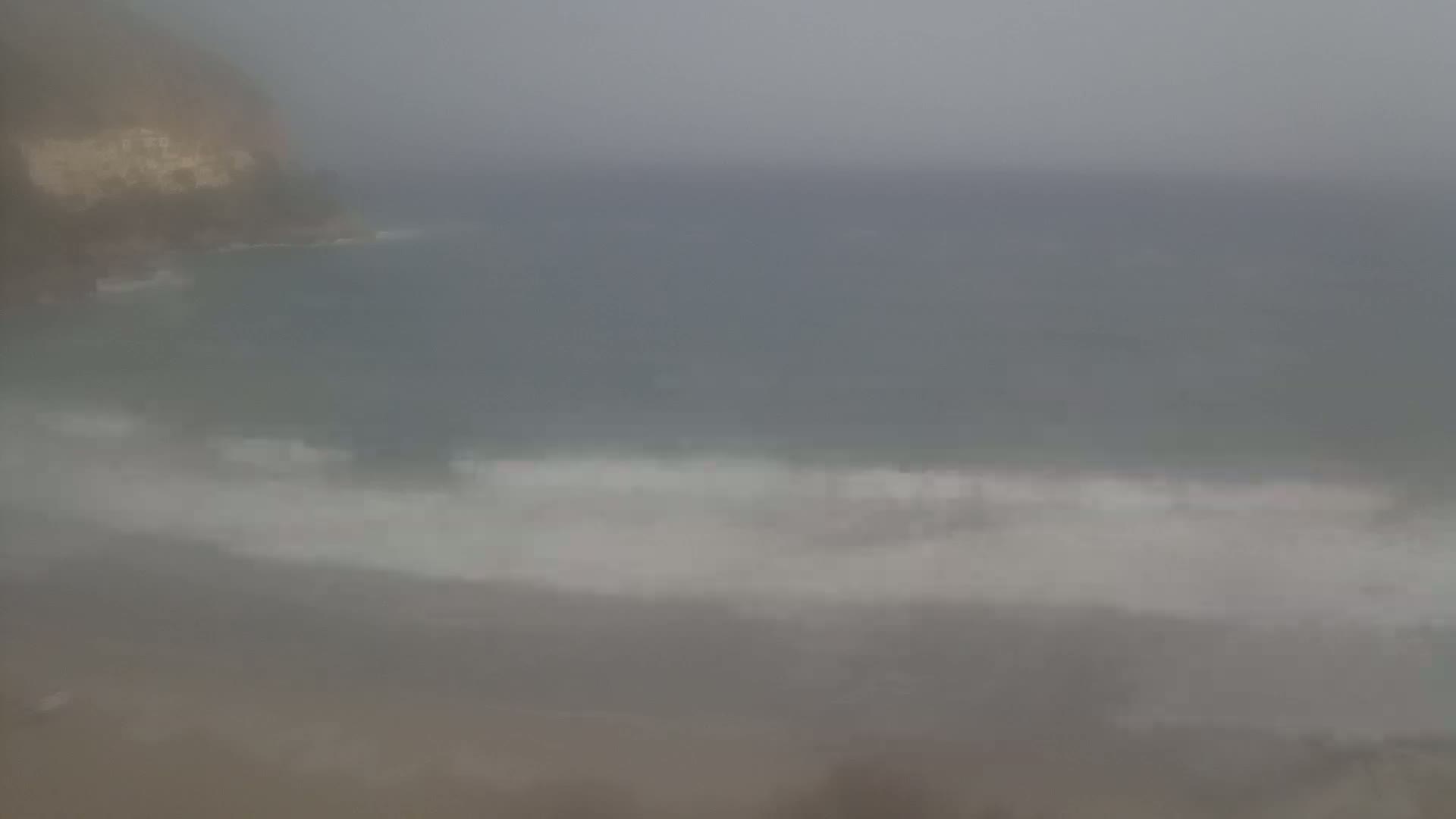 A hazy, overcast day shows a beach with small waves lapping the shore near a cliff face.