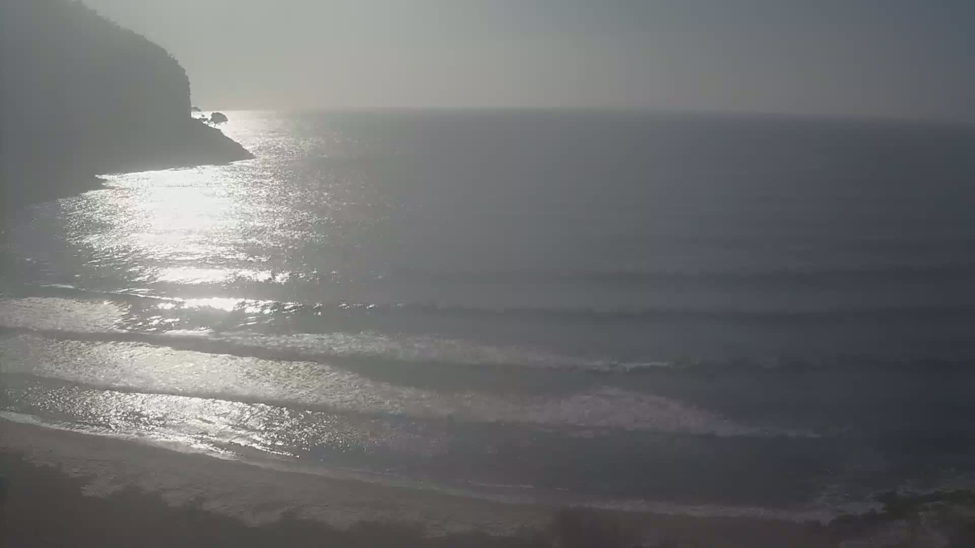 A hazy, sunny day reveals a rocky coastline meeting a calm sea with gentle waves lapping at a sandy beach.