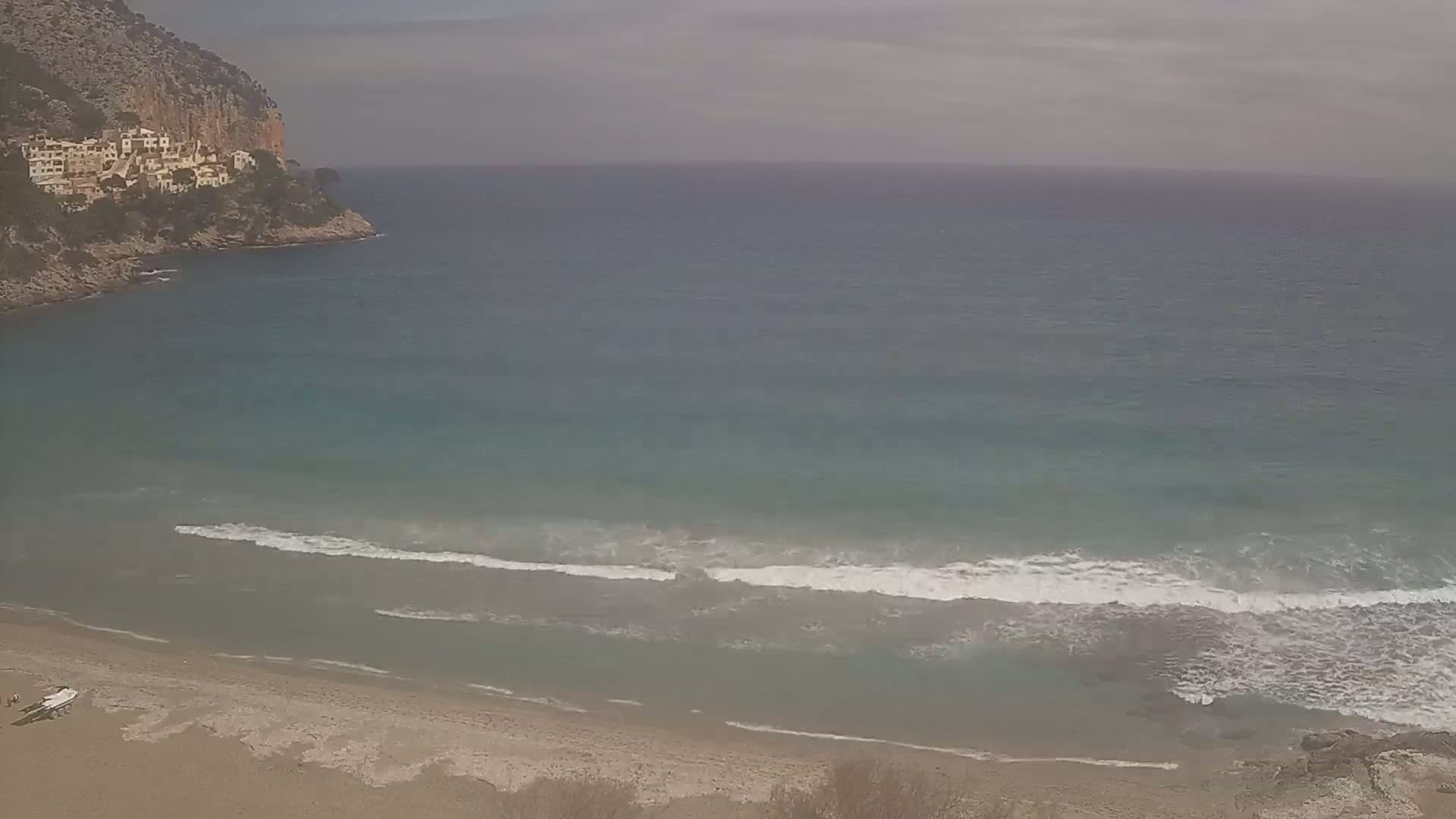 A sandy beach with small waves on a calm, overcast day, next to a rocky cliff with buildings on top.