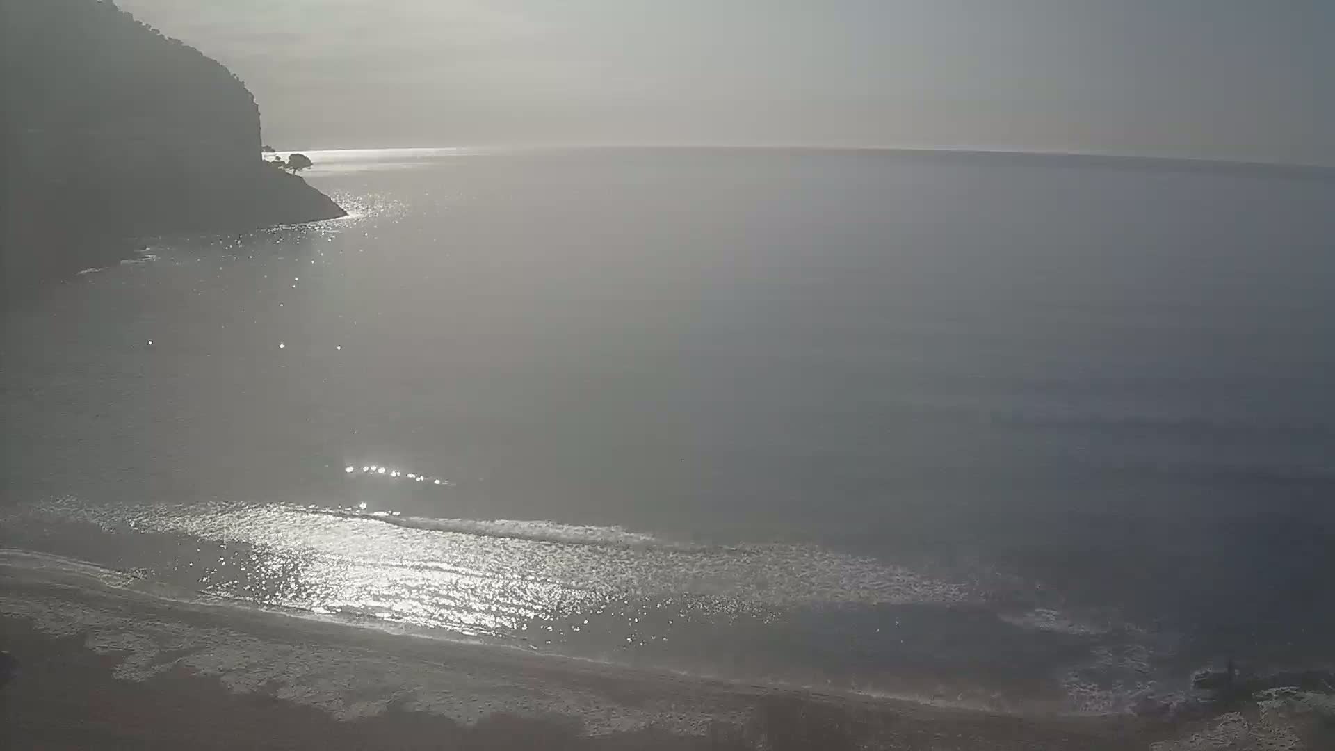 A hazy, sunlit beach scene shows a calm sea lapping a sandy shore near a dark cliff.